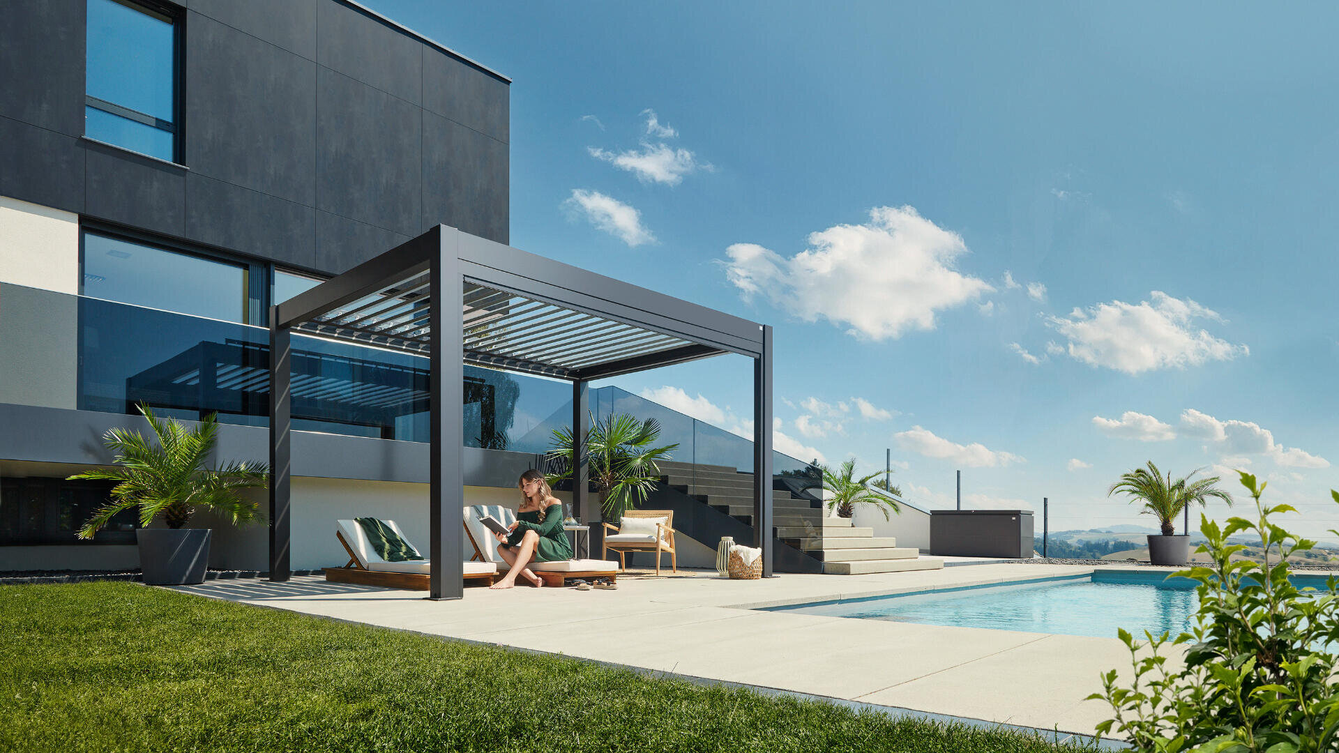 Eine Frau sitzt auf einem Liegestuhl unter einer modernen Pergola neben einem Swimmingpool, mit einem modernen Haus, grünem Rasen, Palmen und einem klaren blauen Himmel im Hintergrund.