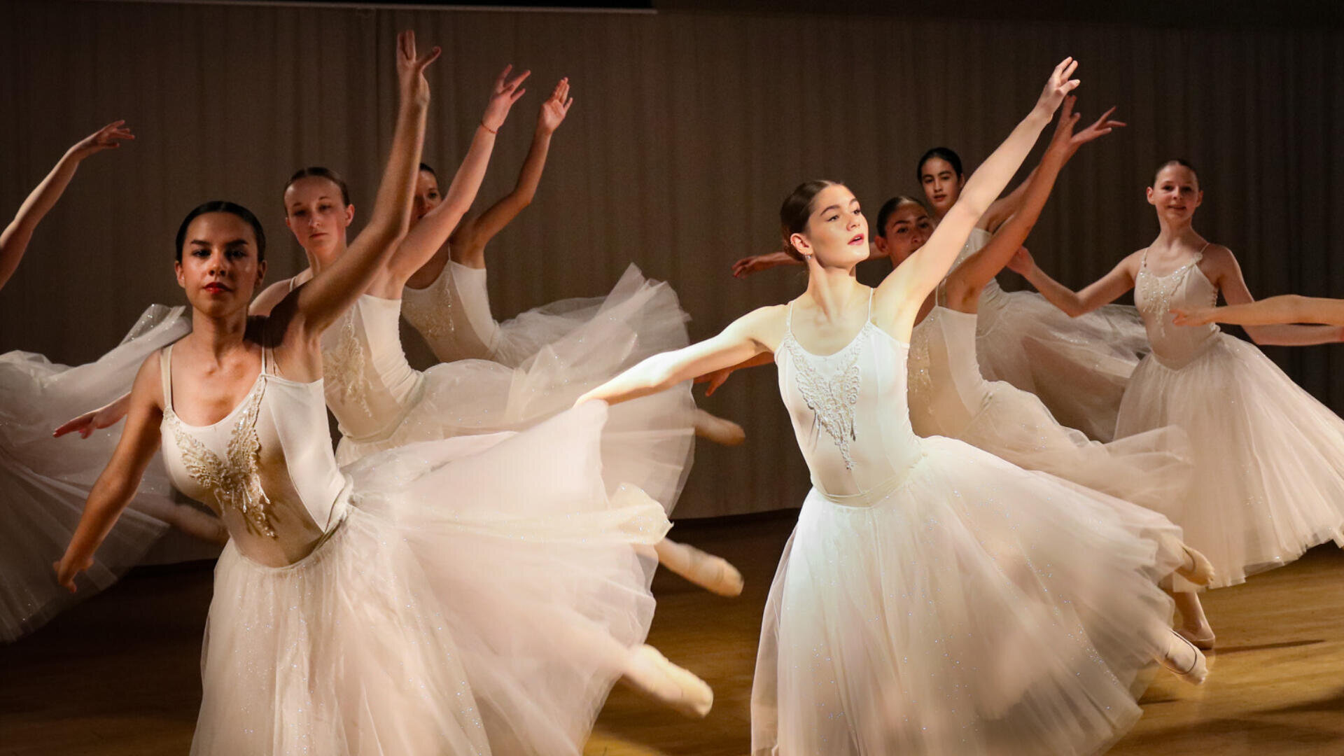Eine Gruppe von Ballerinas in weißen Tutus tanzt anmutig auf der Bühne, mit erhobenen Armen und ausgestreckten Beinen, und fängt einen Moment eleganter Bewegung und Koordination im warmen Bühnenlicht ein.