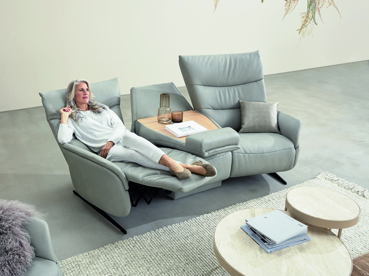 Eine entspannte ältere Frau lümmelt auf einem modernen hellgrauen Liegesofa mit einem dazugehörigen hölzernen Beistelltisch, weichen Kissen und minimalistischem Dekor in einem hellen, modernen Wohnzimmer.