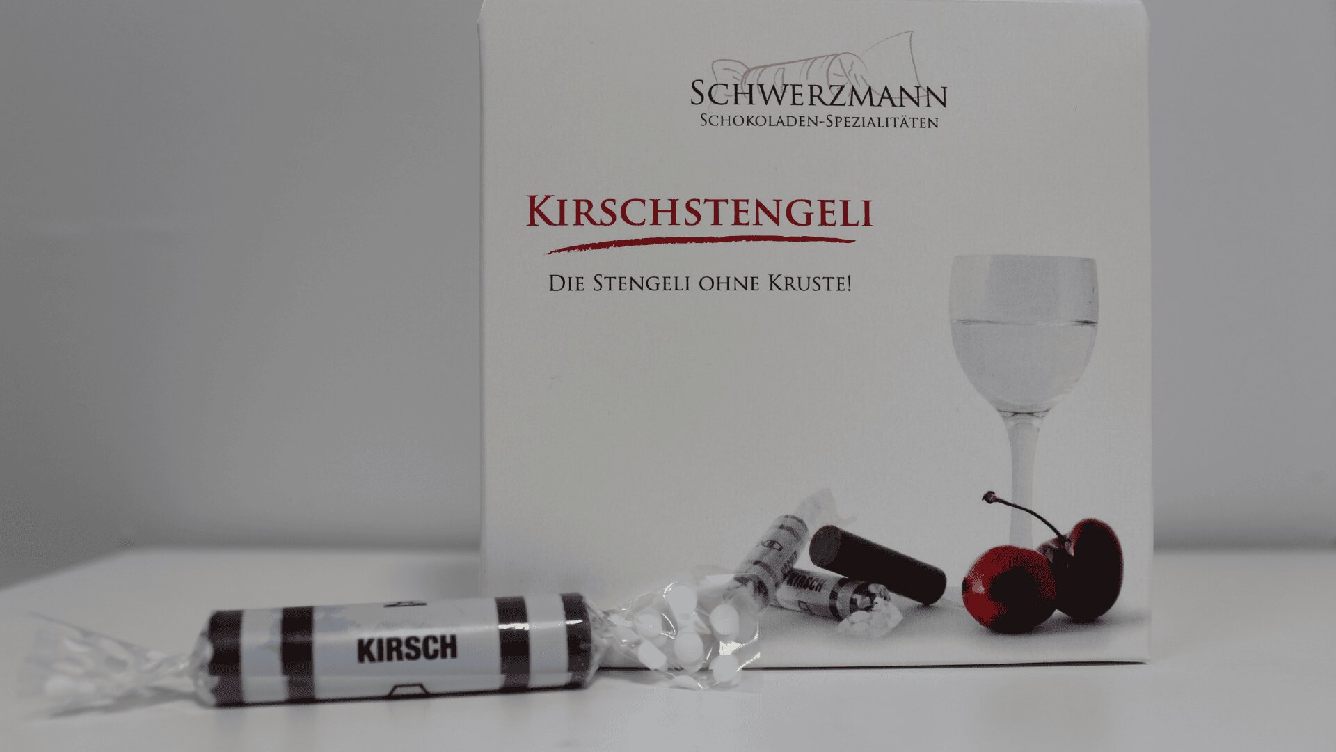 Eine Schachtel mit der Aufschrift Schwerzmann Kirschstengeli mit einem eingewickelten Schokoladenbonbon auf der Vorderseite, einer Abbildung eines Glases, Kirschen und Schokoladenbonbons auf der Schachtel. Der Text lautet: "Die Stengeli ohne Kruste".