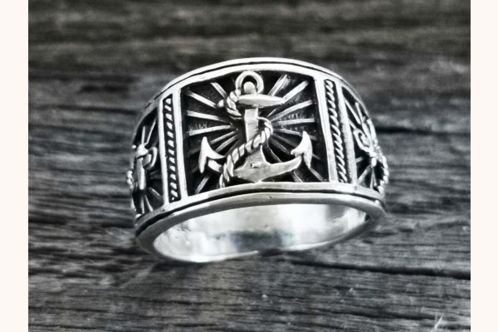 Ein Silberring mit einem erhabenen Ankermotiv in der Mitte, umgeben von Sonnenstrahlmustern, Seildetails und dekorativen Gravuren, präsentiert auf einer rustikalen Holzoberfläche.