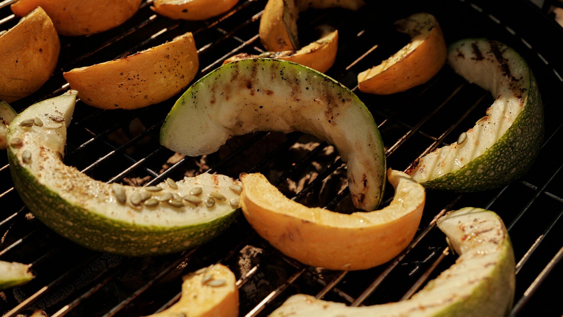 Kürbis- und Melonenscheiben werden auf einem Grill gegrillt. Auf einigen Stücken sind Verkohlungsspuren und Kerne zu sehen, mit einer Nahaufnahme der Grillroste und des rauchigen Hintergrunds.