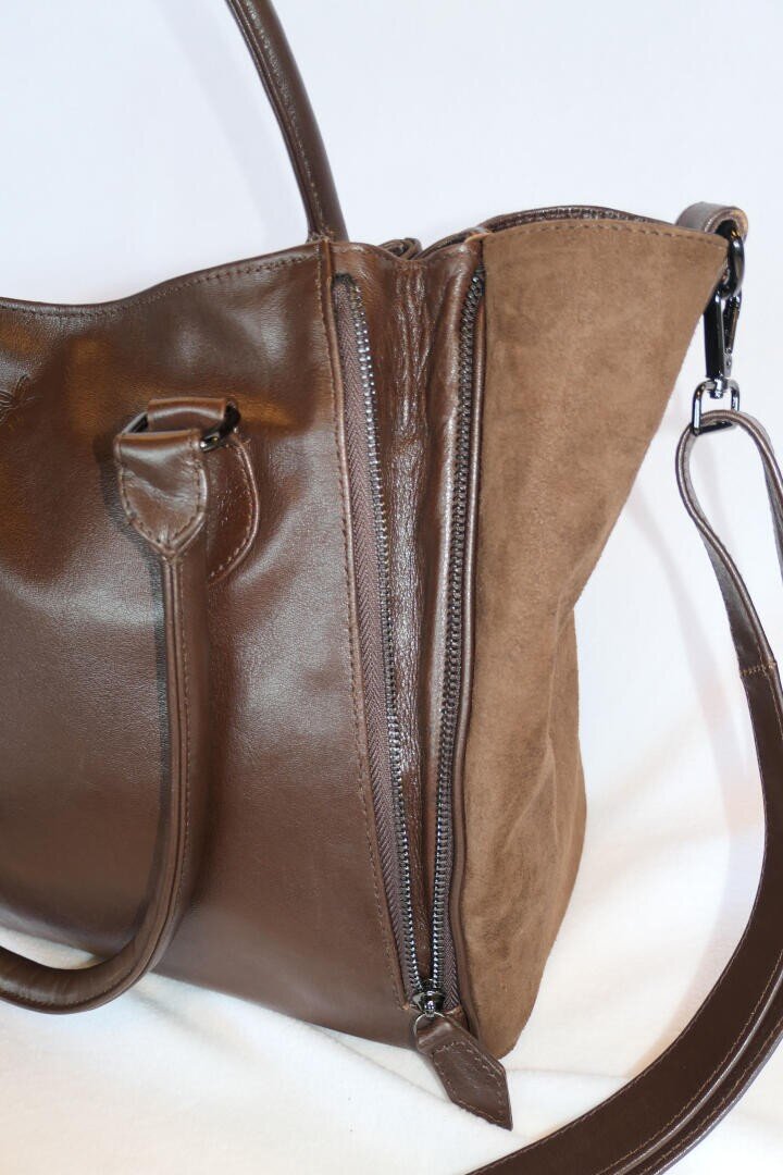 Nahaufnahme einer braunen Leder- und Wildlederhandtasche mit Reißverschluss und verstellbarem Schulterriemen vor einem schlichten weißen Hintergrund.