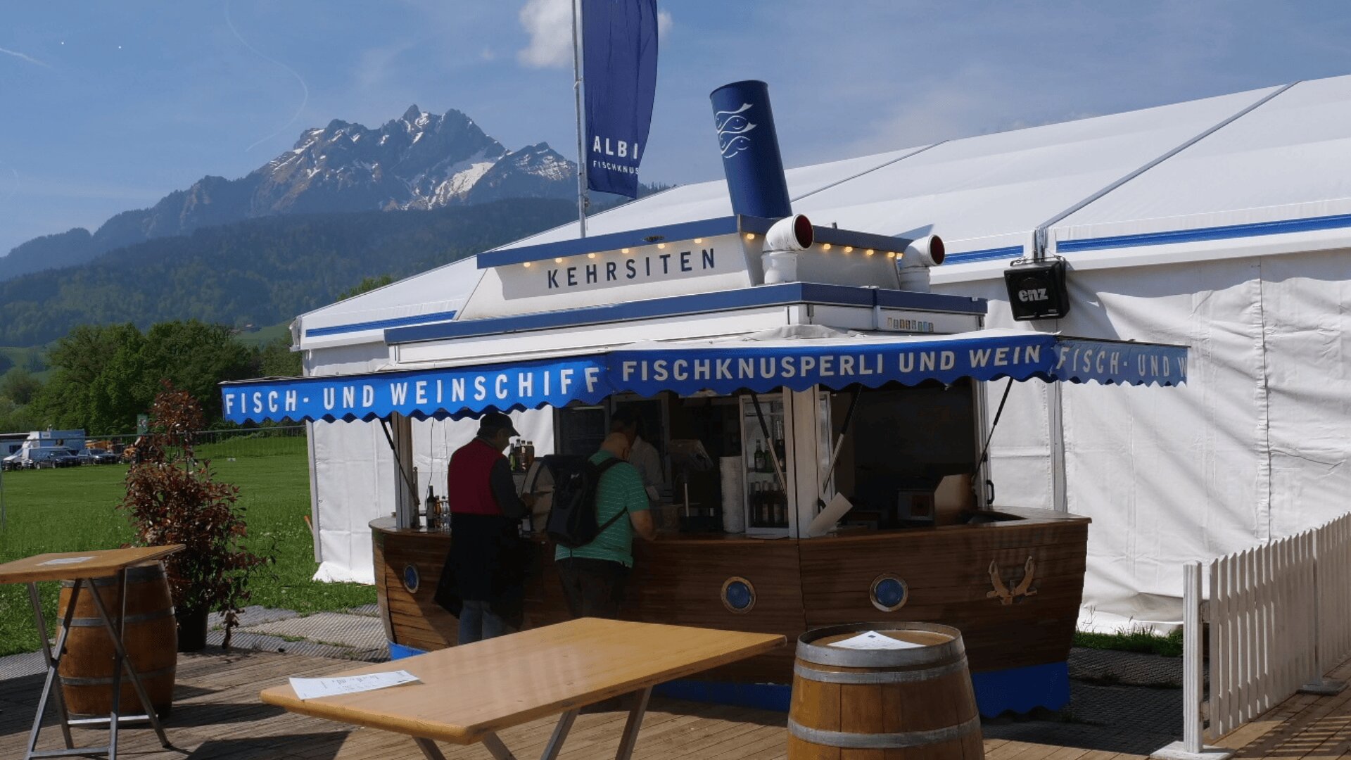 Ein kleiner schiffsförmiger Kiosk mit einer blauen Markise mit der Aufschrift Fisch- und Weinschiff bedient die Kunden im Freien neben einem weißen Zelt, mit Bergen und Grün im Hintergrund.