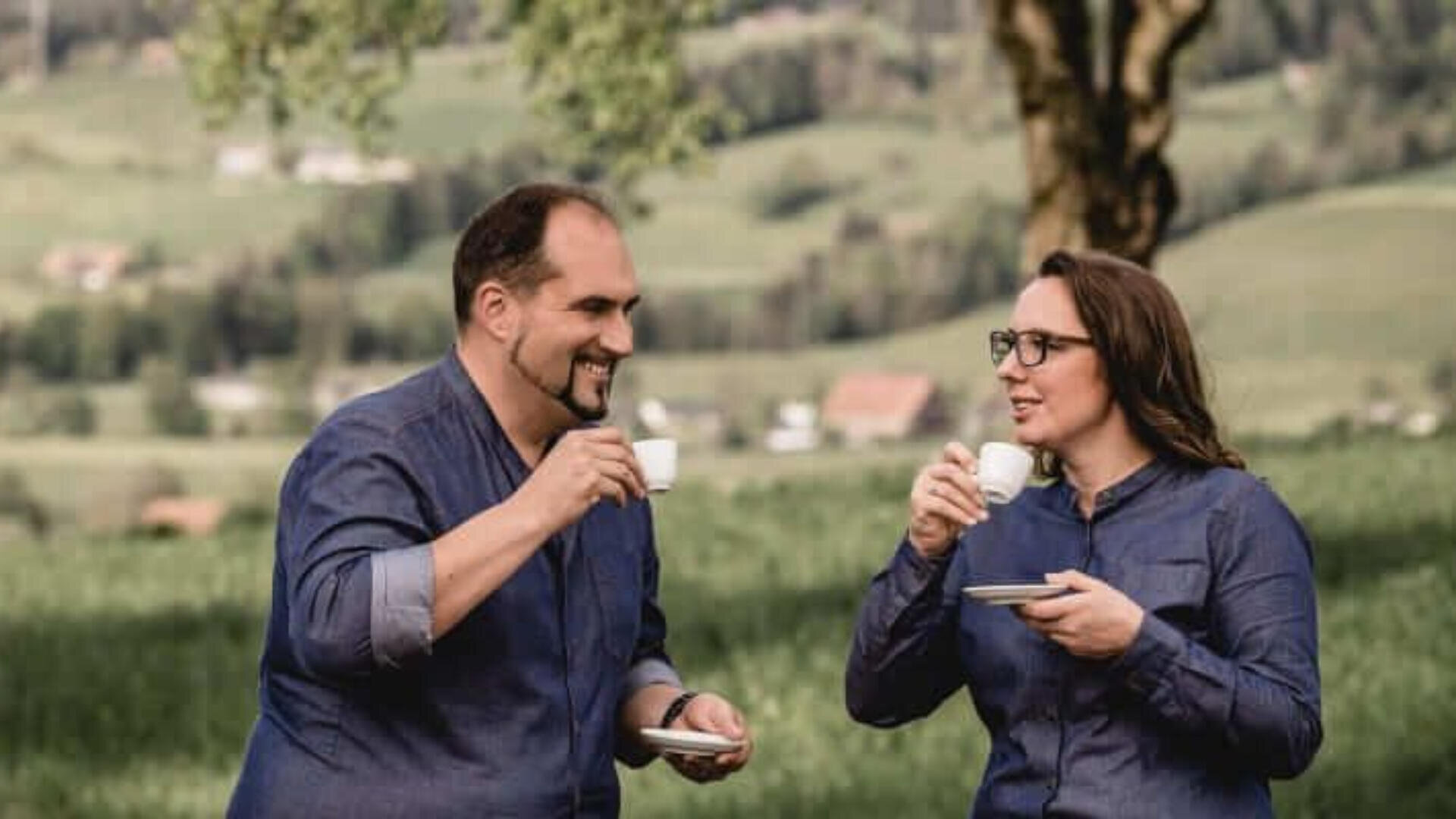 Zwei Personen in passenden Jeanshemden stehen im Freien, lächeln und genießen Kaffee aus kleinen weißen Tassen. Im Hintergrund ist eine verschwommene grüne Landschaft mit Bäumen und fernen Hügeln zu sehen.