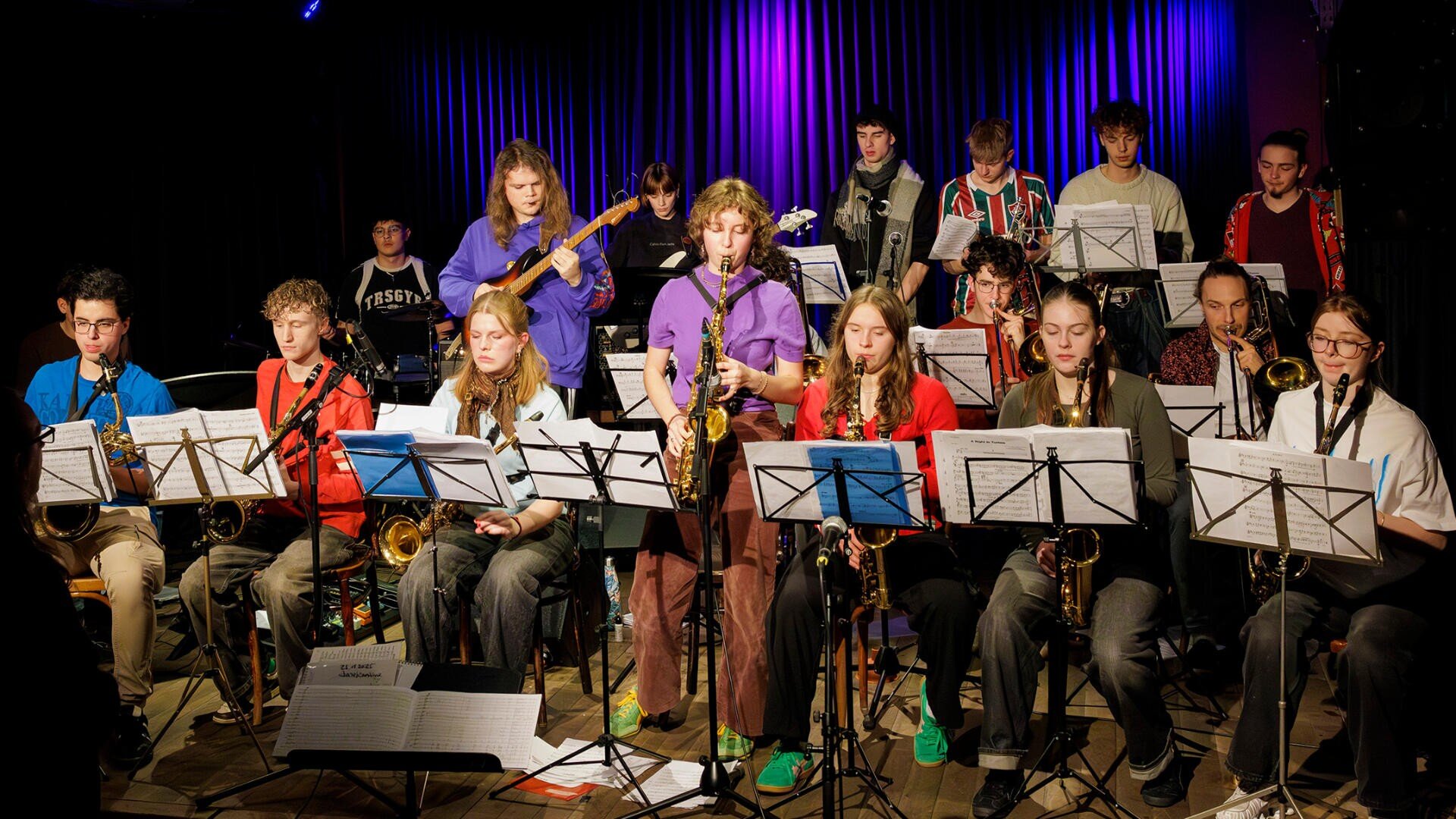 Eine große Jugend-Jazzband tritt auf der Bühne mit Saxophonen, Trompeten, Posaunen, Gitarren und Keyboards auf. Sie sind von Notenständern umgeben und werden von einer violetten Bühnenbeleuchtung angestrahlt.