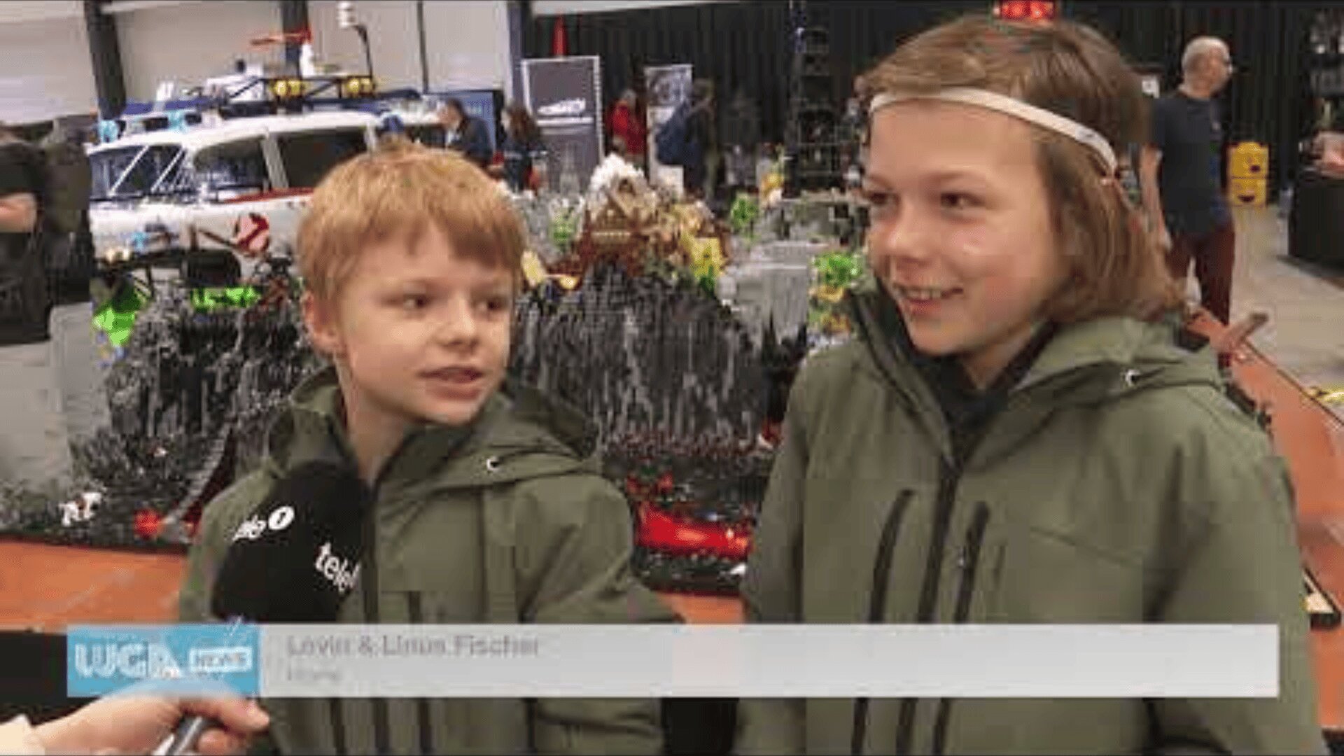 Zwei Kinder in grünen Jacken werden in einer LEGO Ausstellung interviewt. Sie stehen vor einer großen, detaillierten LEGO Ausstellung, und einer spricht in ein Mikrofon mit der Aufschrift tele.