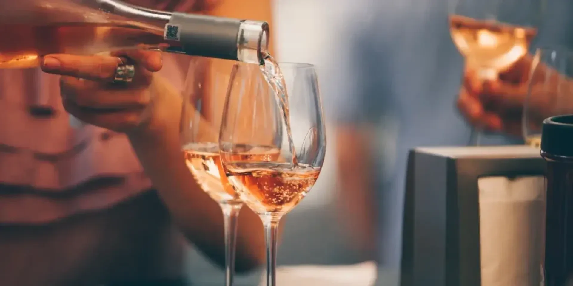Eine Person gießt Roséwein aus einer Flasche in ein Glas ein, während zwei andere Weingläser in der Nähe teilweise gefüllt sind. Eine andere Hand hält ein Glas im Hintergrund. Es scheint sich um ein geselliges Beisammensein zu handeln.