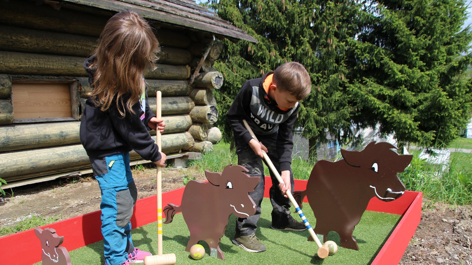 Zwei Kinder spielen im Freien Minigolf, indem sie Bälle mit Schlägern durch ausgeschnittene Kuhfiguren auf einem kleinen Kunstrasenplatz neben einer Blockhütte schlagen, mit Bäumen im Hintergrund.