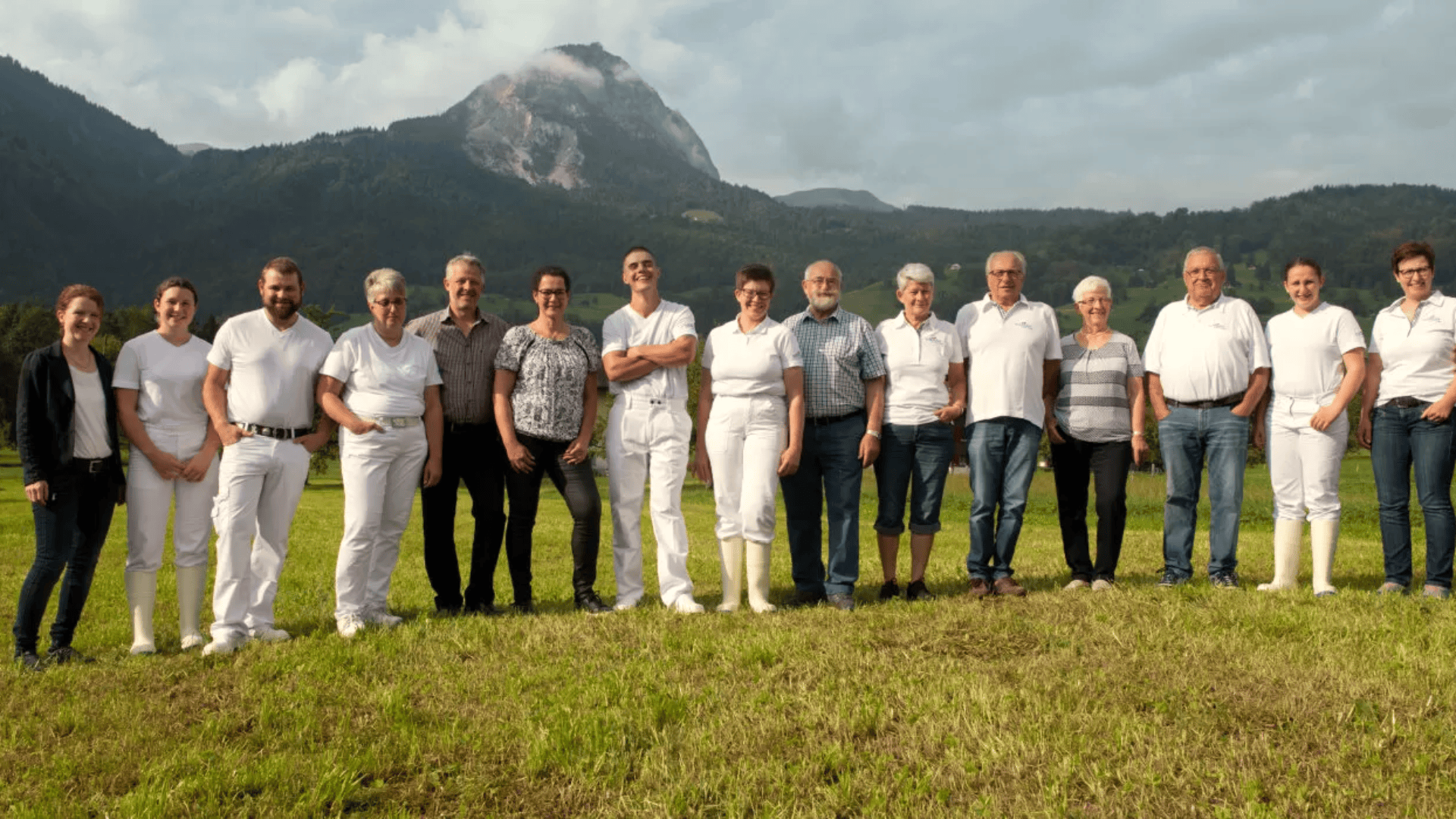 Eine Gruppe von fünfzehn Personen steht auf einer grasbewachsenen Wiese mit Bergen im Hintergrund. Die meisten tragen weiße Kleidung und Stiefel, einige wenige tragen Freizeitkleidung. Der Himmel ist bewölkt.