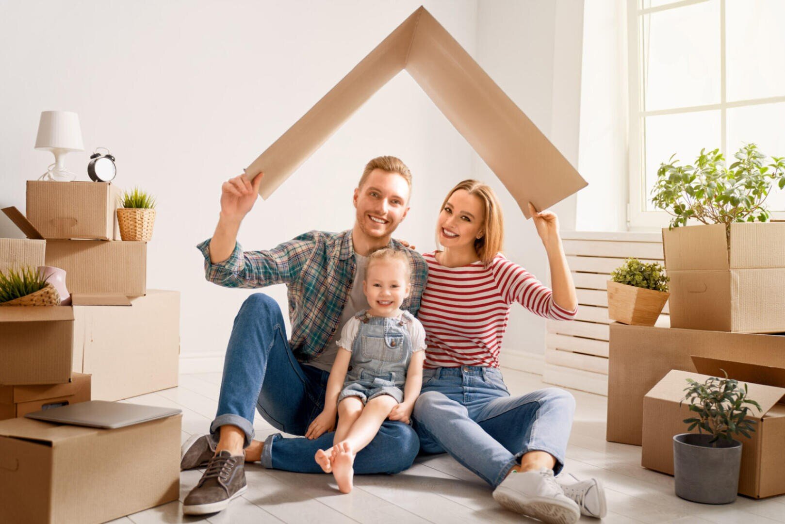 Eine lächelnde dreiköpfige Familie sitzt zwischen Umzugskartons auf dem Boden und hält eine Dachform aus Pappe über ihren Köpfen, die ein neues Zuhause symbolisiert. Sonnenlicht strömt durch ein Fenster in den hellen Raum.