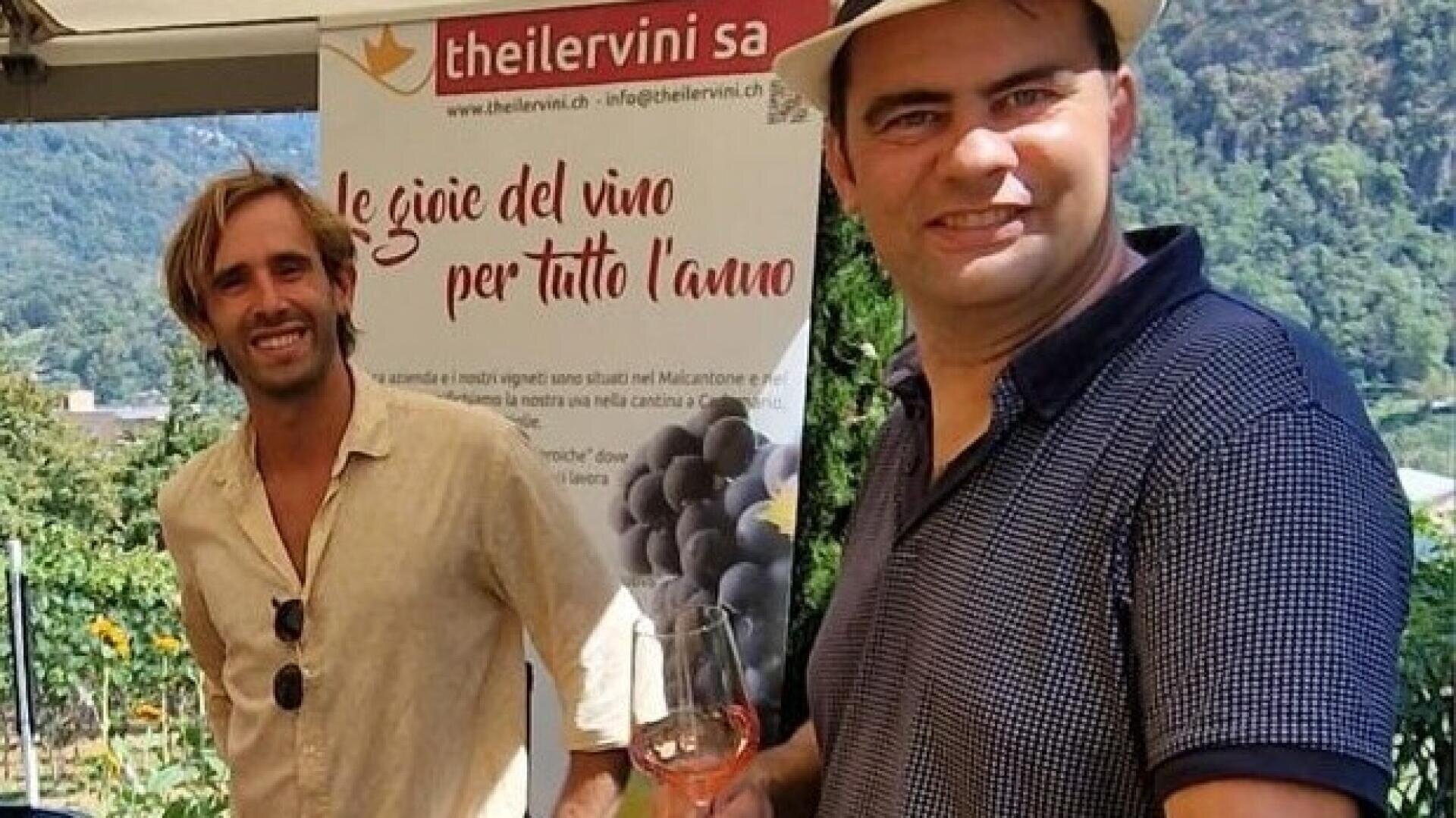 Zwei Männer stehen bei einer Weinverkostung im Freien. Einer trägt ein beiges Hemd, der andere ein marineblaues Hemd und einen Hut und hält ein Weinglas in der Hand. Hinter ihnen ist ein Banner mit Trauben und italienischem Text zu sehen, und im Hintergrund sind Berge zu sehen.