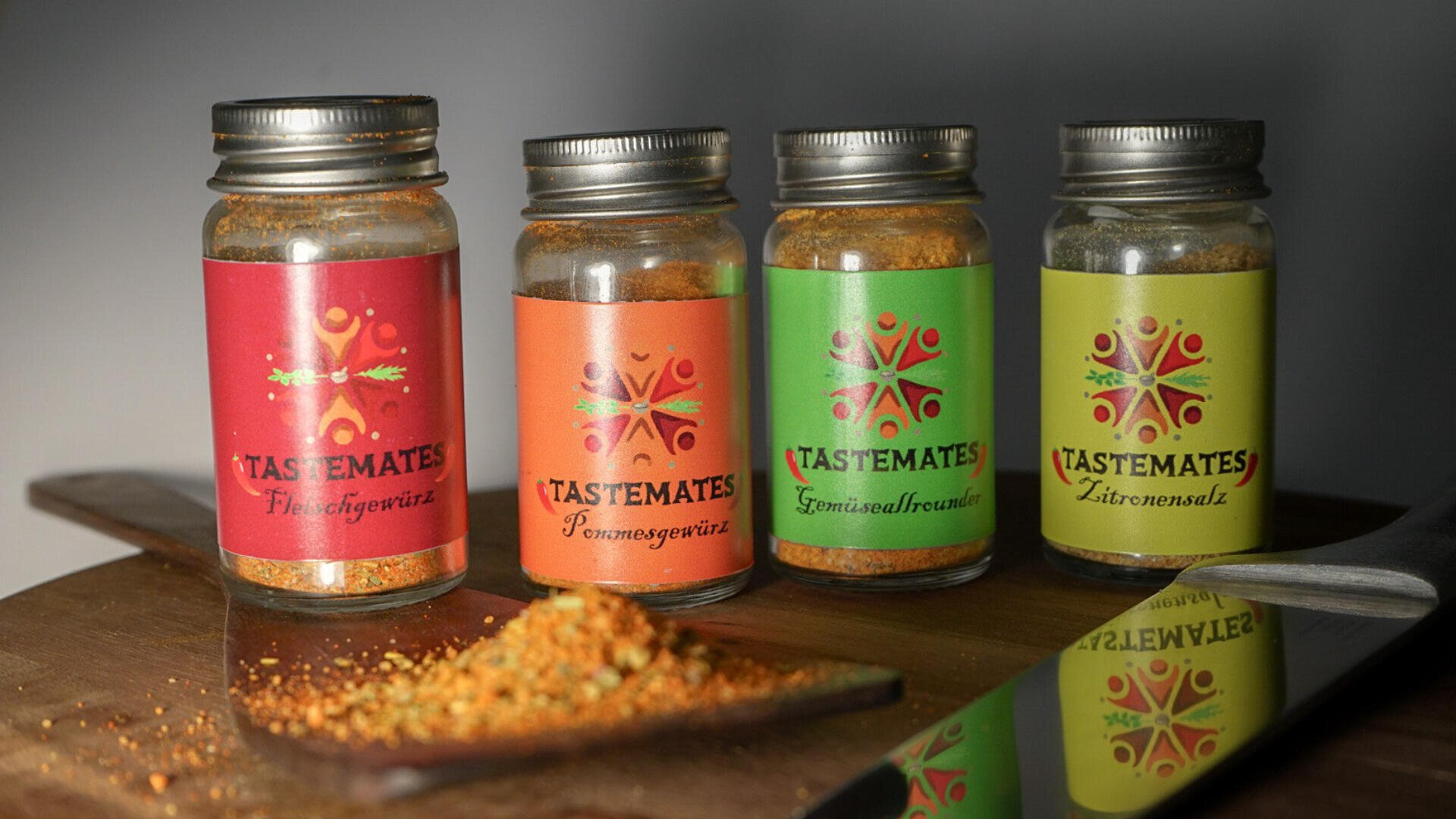 Vier Gewürzdosen mit bunten Etiketten (rot, orange, grün, gelb) mit der Aufschrift TASTEMATES stehen auf einer hölzernen Unterlage, vor denen etwas Gewürzpulver verschüttet ist, und ein Messer liegt daneben.
