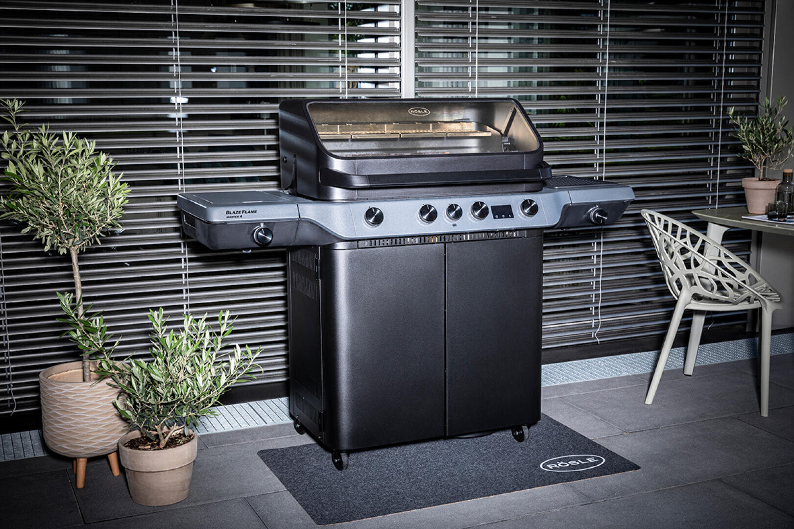 Ein großer schwarzer Gasgrill mit geschlossenem Deckel steht auf einem Balkon, umgeben von Topfpflanzen und Gartenmöbeln. Im Hintergrund sind horizontale Jalousien zu sehen.