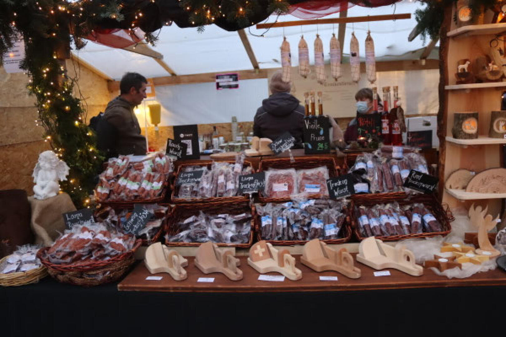 Ein mit Lichtern und Grünzeug geschmückter Marktstand bietet verschiedene Wurstsorten in Körben, hölzerne Schweizer Souvenirs und Hängewürste an. Zwei Verkäuferinnen stehen hinter der Theke und bedienen die Kunden.