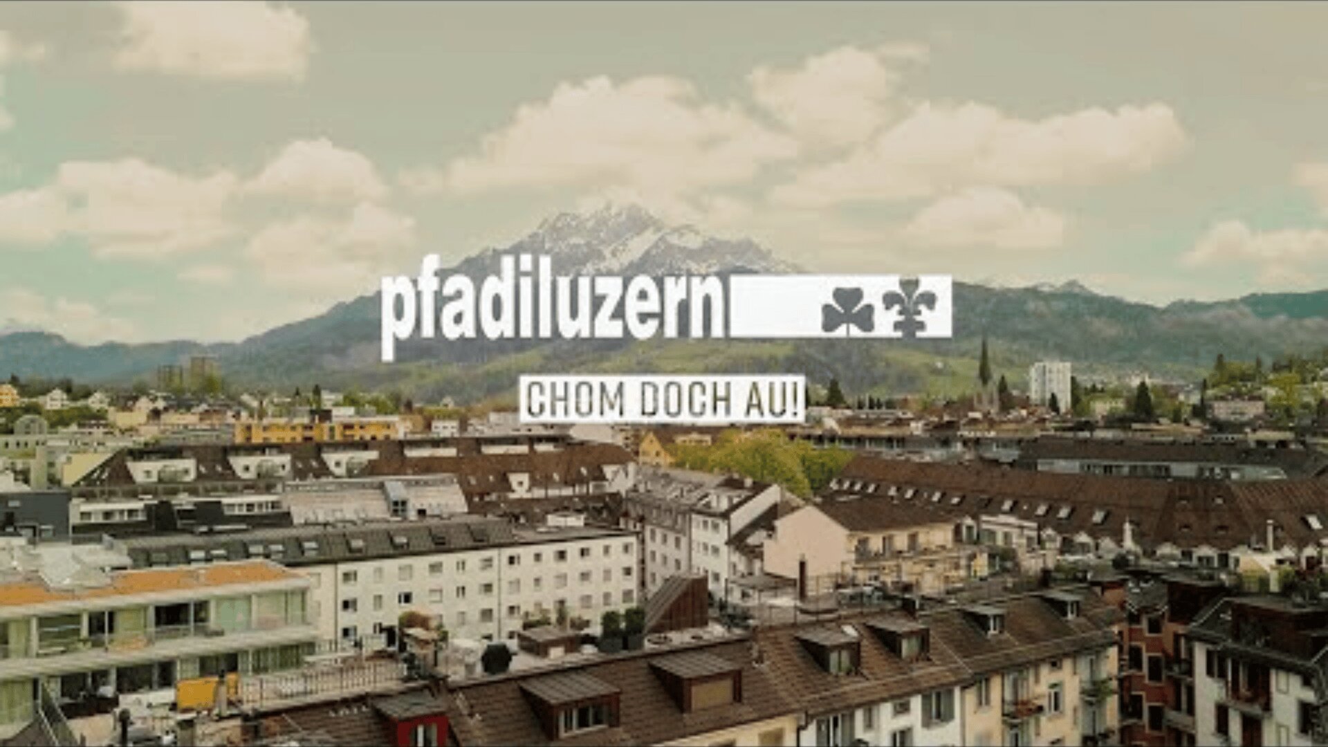 Luftaufnahme einer Stadt mit Häuserzeilen und einem Berg im Hintergrund. Überlagert wird der Text "Pfadiluzern" mit zwei Kleesymbolen und darunter "CHOM DOCH AU!" in einem rechteckigen Kasten.
