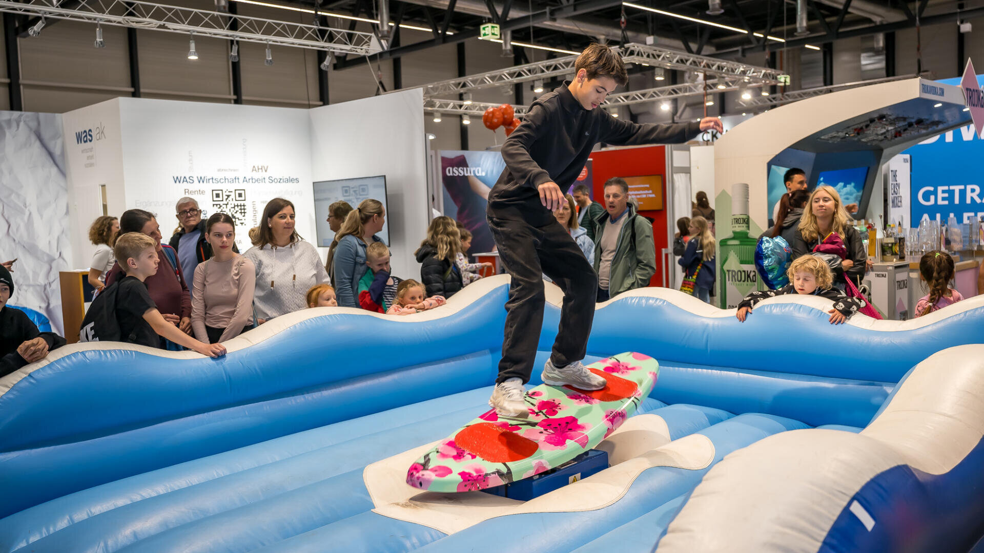 Ein junger Mensch balanciert auf einem bunten mechanischen Surfbrett, umgeben von aufblasbaren blauen Wellen, während eine Menschenmenge, darunter Kinder und Erwachsene, bei einer Indoor-Veranstaltung oder Ausstellung aufmerksam zusieht.