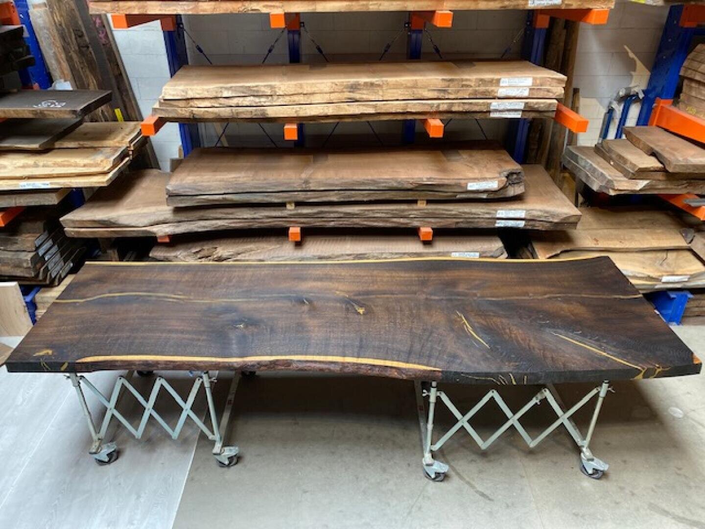 Eine große, dunkle Holzplatte mit einer natürlichen, unebenen Kante ruht auf zwei Metallständern mit Rädern. Dahinter sind mehrere andere Holzplatten auf blauen und orangefarbenen Regalen in einer Werkstatt gestapelt.