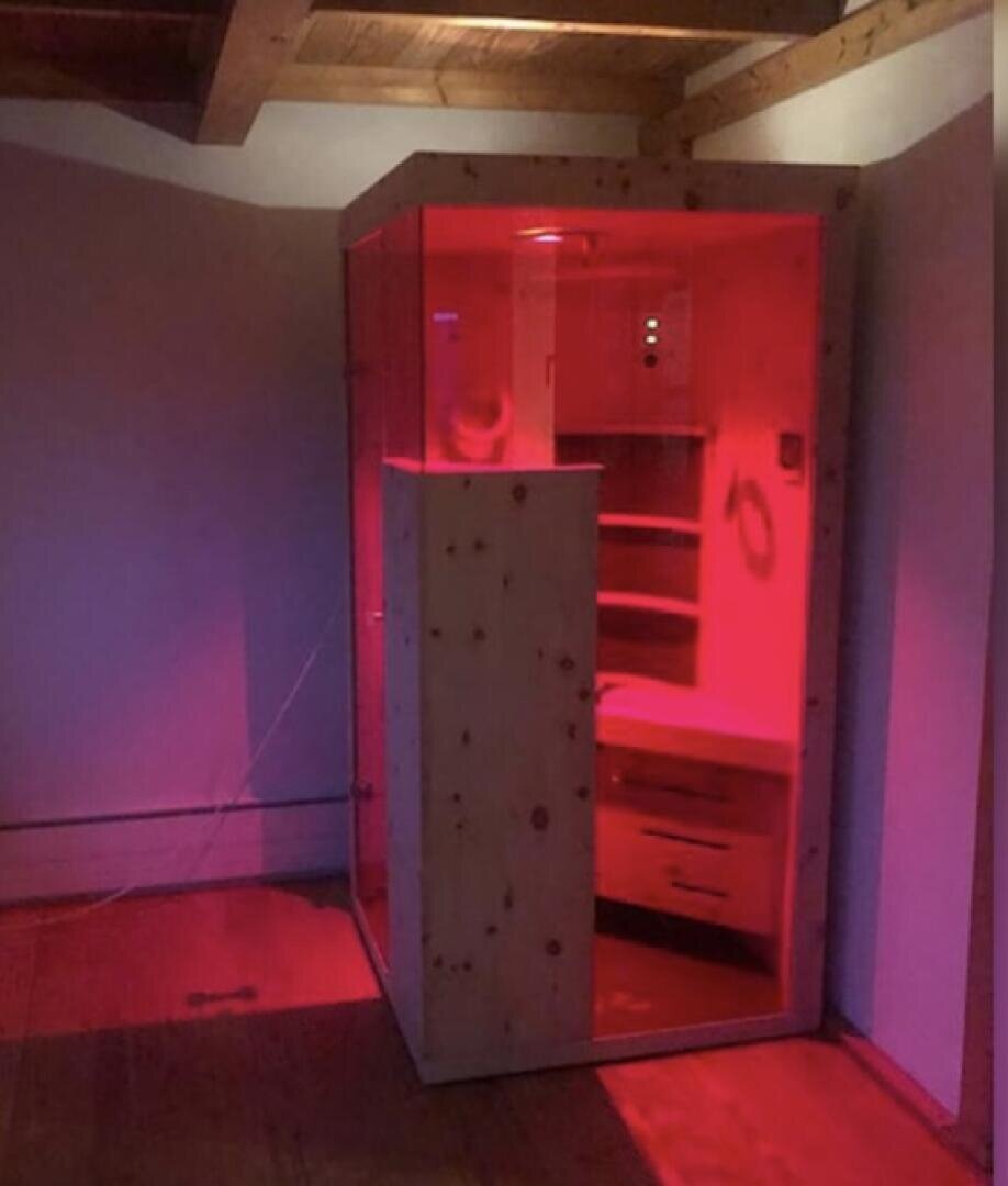 Eine Holzsauna oder ein Dampfbad mit roter Beleuchtung steht in der Ecke eines Raums mit Holzböden und Deckenbalken. Die Sauna hat eine Glastür und Regale im Inneren, die ein warmes rotes Licht ausstrahlen.