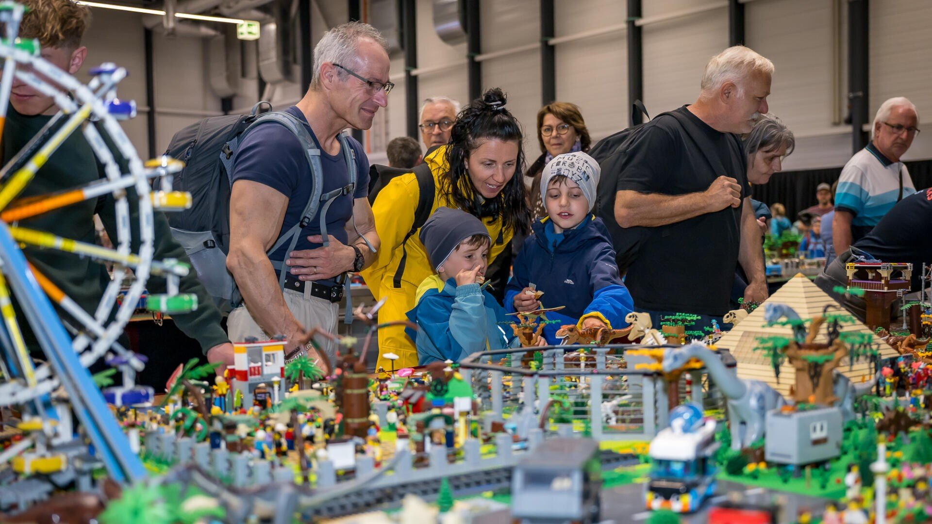 Eine Gruppe von Erwachsenen und Kindern versammelt sich um eine große, farbenfrohe LEGO-Ausstellung mit einem Riesenrad, Gebäuden und kleinen Figuren und untersucht die Details bei einer Indoor-Veranstaltung oder Ausstellung.