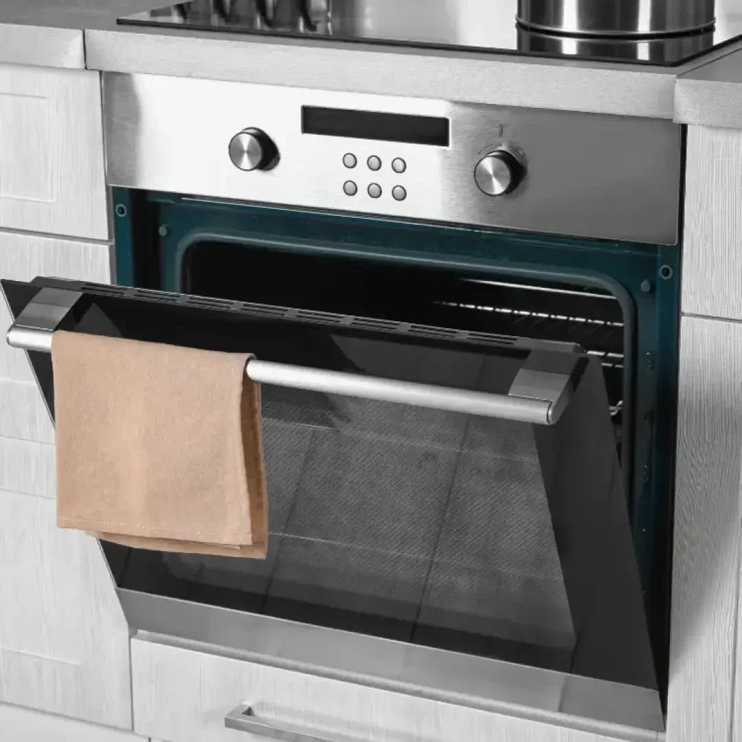 Ein moderner Einbaubackofen aus rostfreiem Stahl mit offener Tür und einem beigen Küchentuch am Griff, eingebettet in helle Holzschränke.