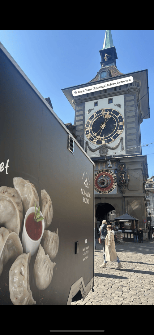 Ein Foodtruck mit einem Bild von Knödeln und einer Tasse Soße steht in der Nähe des historischen Zytglogge-Uhrturms in Bern, Schweiz, während die Leute an einem sonnigen Tag vorbeilaufen.