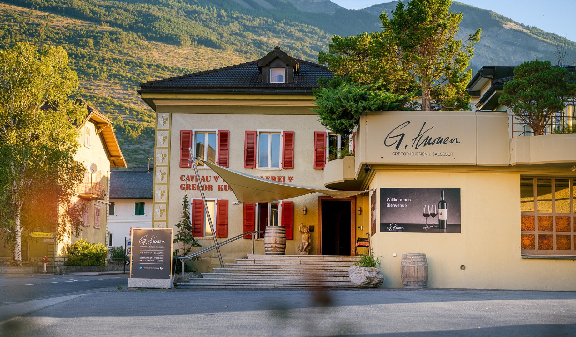 Ein Kellereigebäude mit roten Fensterläden, beigen Wänden und einem Schild mit der Aufschrift G. Kuonen in einer Berglandschaft. Am Eingang unter einem Sonnenschirm sind Weinfässer, Pflanzen und Willkommensschilder zu sehen.