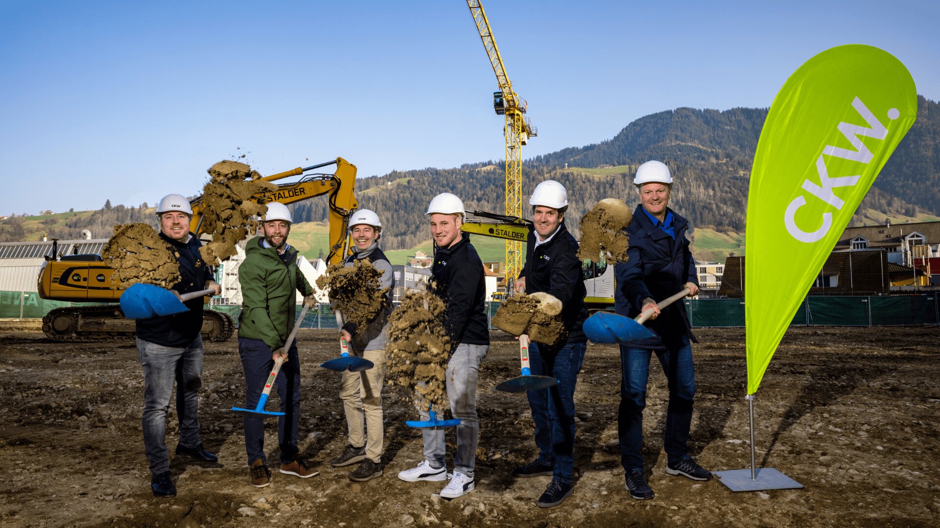 Sechs Personen mit Helmen halten Schaufeln und schaufeln Erde auf einer Baustelle mit Baggern und einem Kran im Hintergrund. Rechts steht eine grüne CKW-Flagge, in der Ferne sind Hügel und blauer Himmel zu sehen.