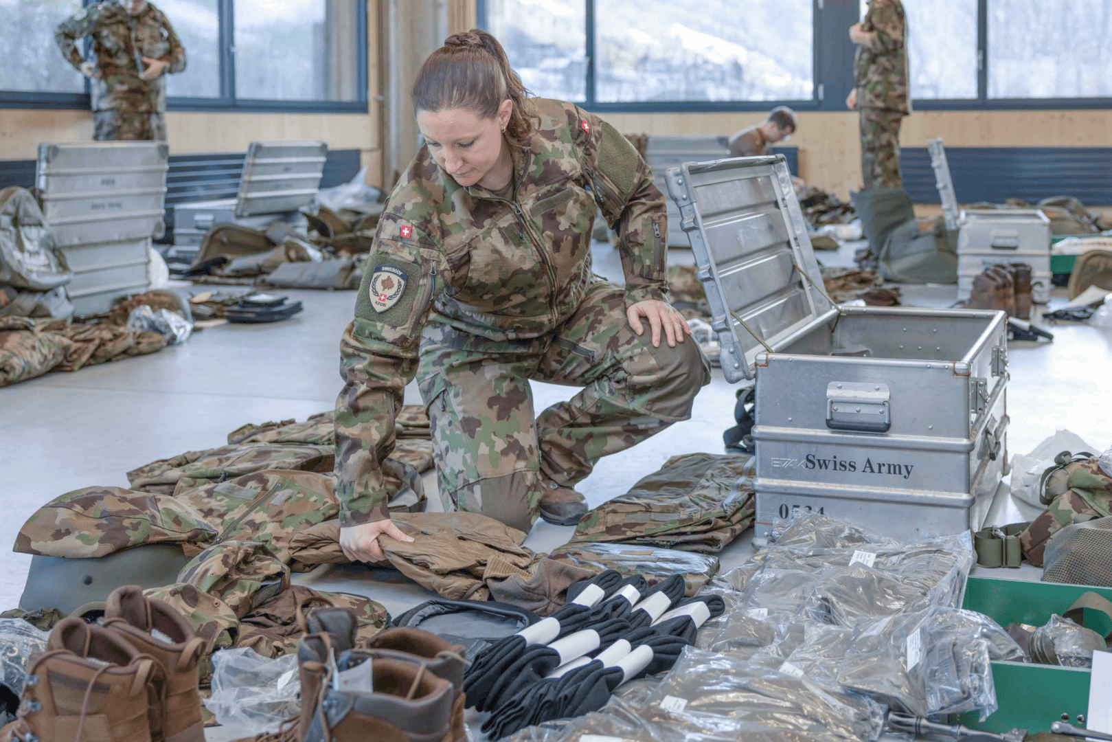 Eine Frau in Schweizer Armeeuniform kniet auf dem Boden und sortiert in einem großen, hellen Raum mit mehreren anderen Soldaten im Hintergrund militärische Ausrüstung und Kleidung zwischen offenen Ausrüstungskoffern.