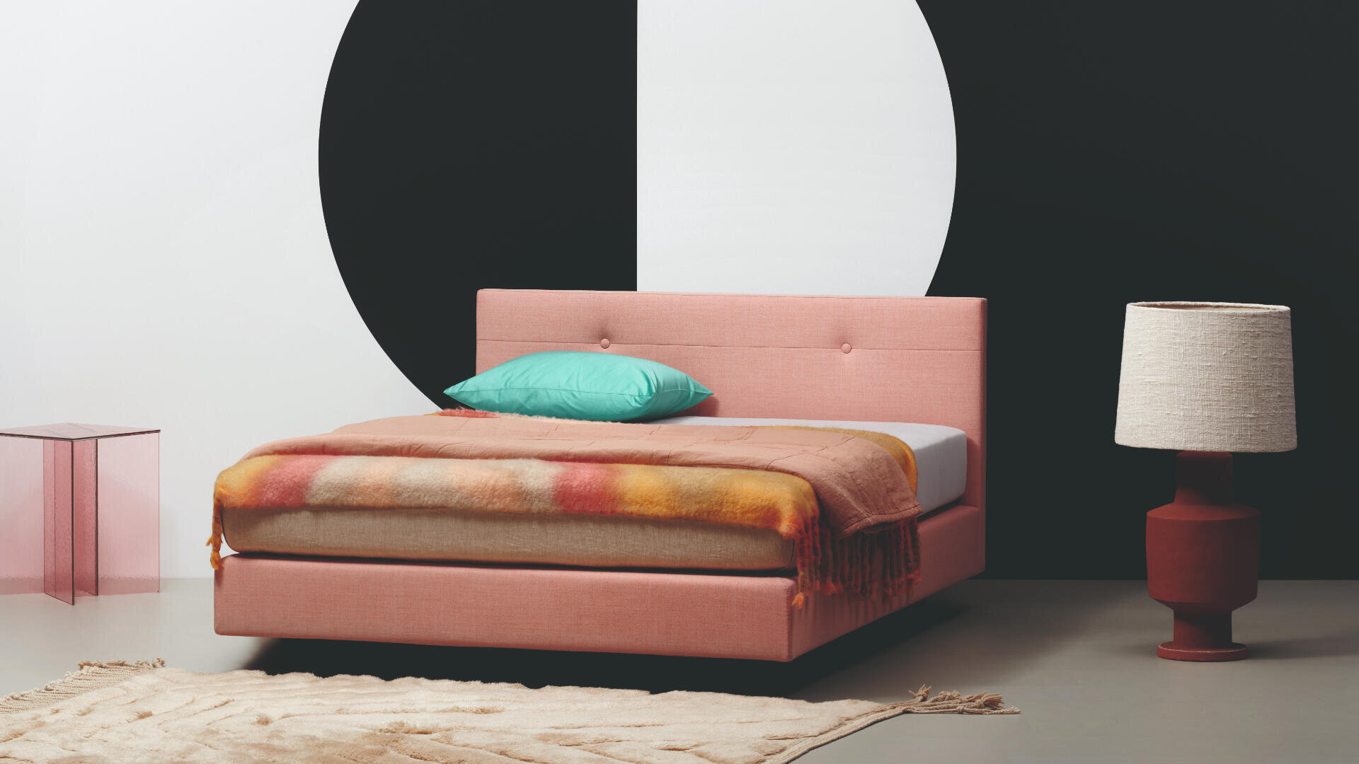 Ein modernes Schlafzimmer mit einem rosa gepolsterten Bett, einer bunten Decke, einem blaugrünen Kissen, großer geometrischer Wandkunst in Weiß und Schwarz, einer roten Lampe mit beigem Schirm, einem rosa Beistelltisch und einem cremefarbenen Teppich mit Struktur.
