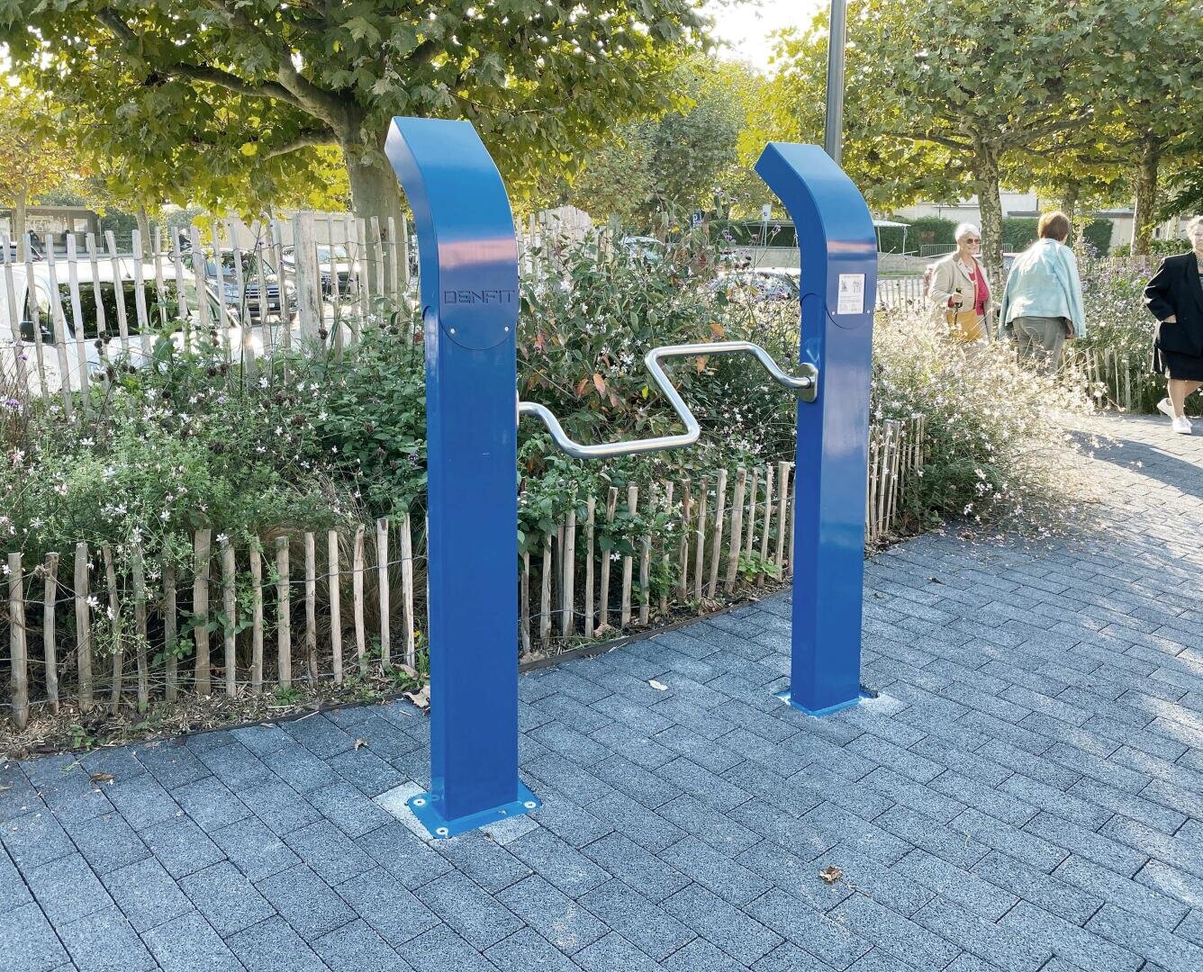 Eine blaue Outdoor-Trainingsstation mit zwei aufrechten Pfosten, die durch eine zickzackförmige Metallstange verbunden sind, ist auf einem gepflasterten Gehweg in der Nähe eines Gartens mit Blumen und Bäumen installiert; die Menschen gehen in der Nähe spazieren.