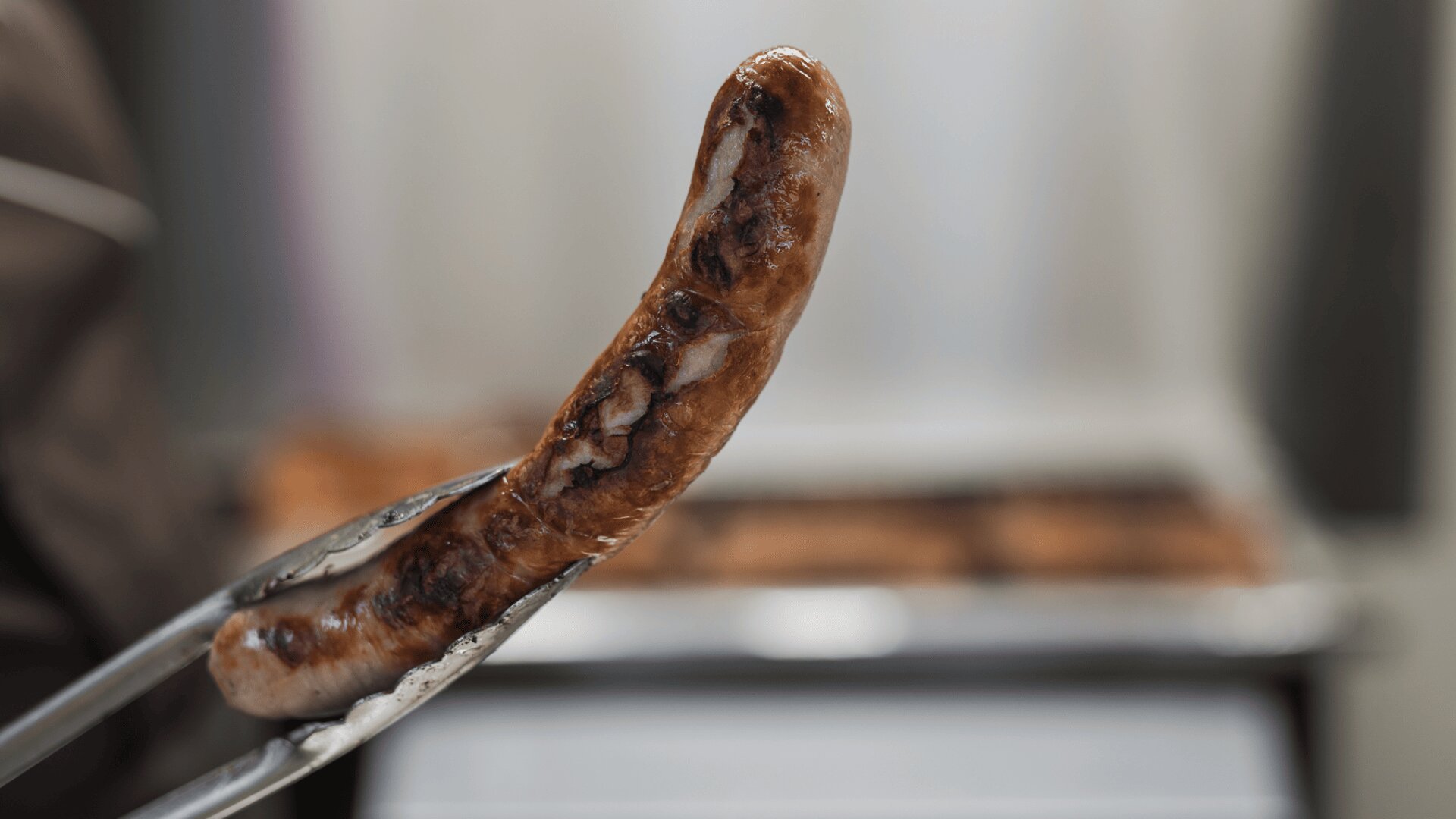 Eine Metallzange hält eine gegrillte Wurst, deren Oberfläche gebräunt und leicht verkohlt ist, mit unscharfem Hintergrund.