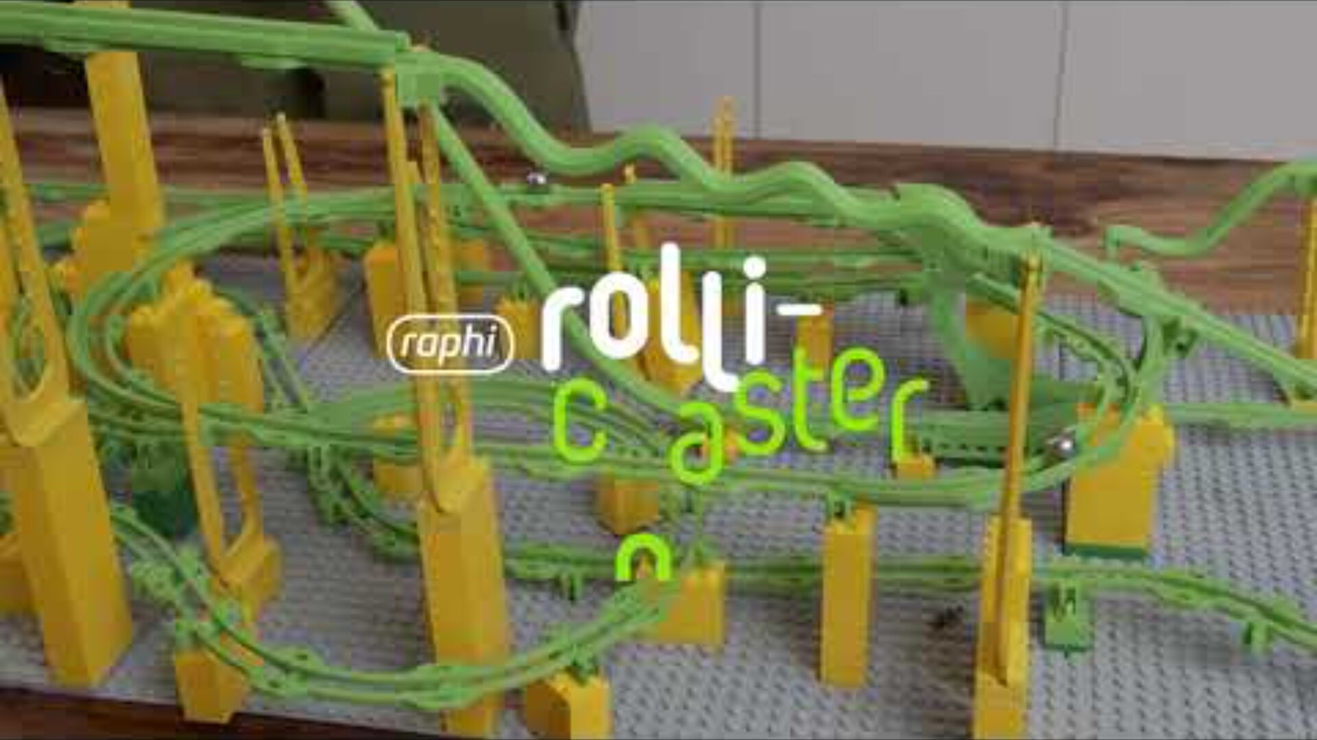 Ein Spielzeug-Achterbahnset mit leuchtend grünen Schienen und gelben Stützen steht auf einem Tisch. Der Text über dem Bild lautet raphi rolli-coaster.