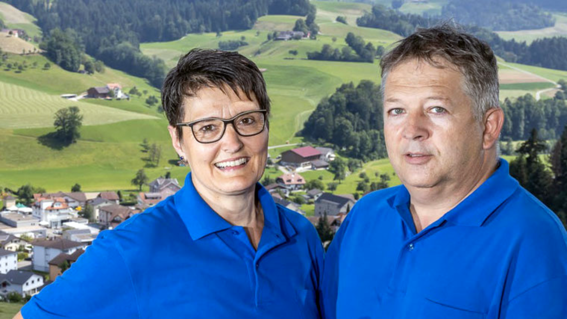 Zwei Erwachsene in blauen Polohemden stehen lächelnd nebeneinander, mit einer malerischen ländlichen Landschaft aus grünen Feldern, Bäumen und kleinen Gebäuden im Hintergrund.