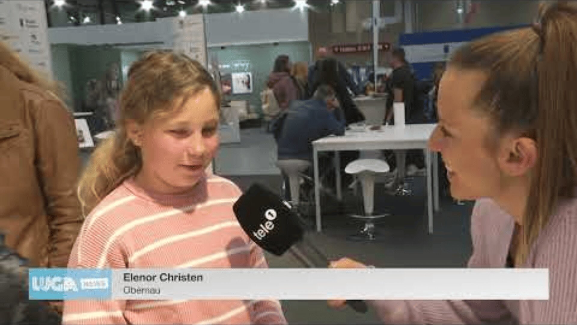 Ein Mädchen in einem rosa gestreiften Pullover wird von einer Frau interviewt, die ein Mikrofon mit einem Tele-Logo hält. Im Hintergrund sitzen und gehen Menschen bei einer Veranstaltung, die in einem Gebäude zu sein scheint.