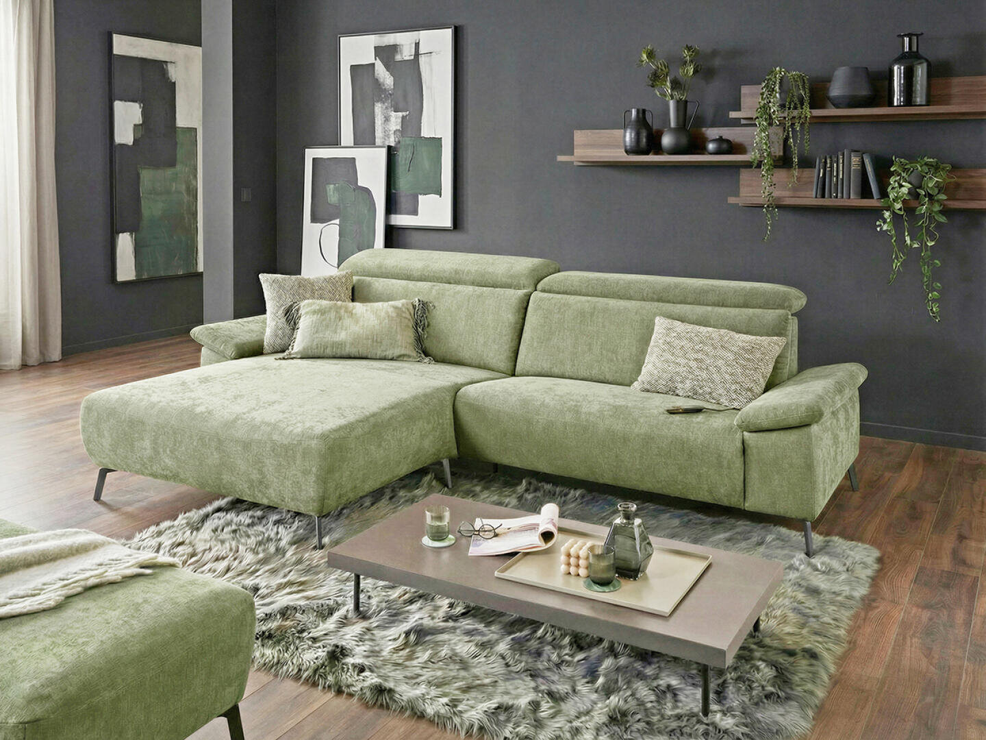 Ein modernes Wohnzimmer mit einem grünen Sektionssofa, abstrakter Kunst an den dunklen Wänden, einem zotteligen Teppich, einem niedrigen Couchtisch mit Büchern und Kerzen und schwebenden Regalen, die mit Pflanzen und Vasen dekoriert sind.