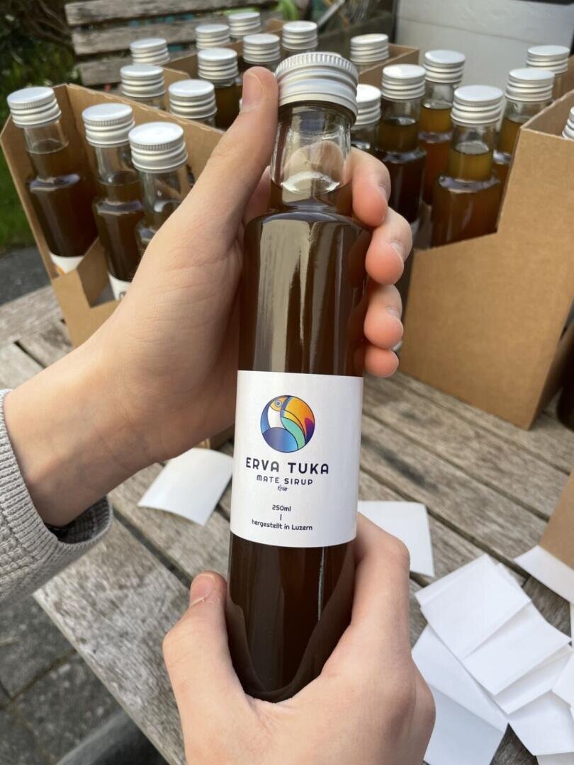 Eine Person hält eine Flasche Erva Tuka Mate Sirup mit einem bunten Vogellogo, umgeben von ähnlichen Flaschen in Pappträgern auf einem Holztisch im Freien.