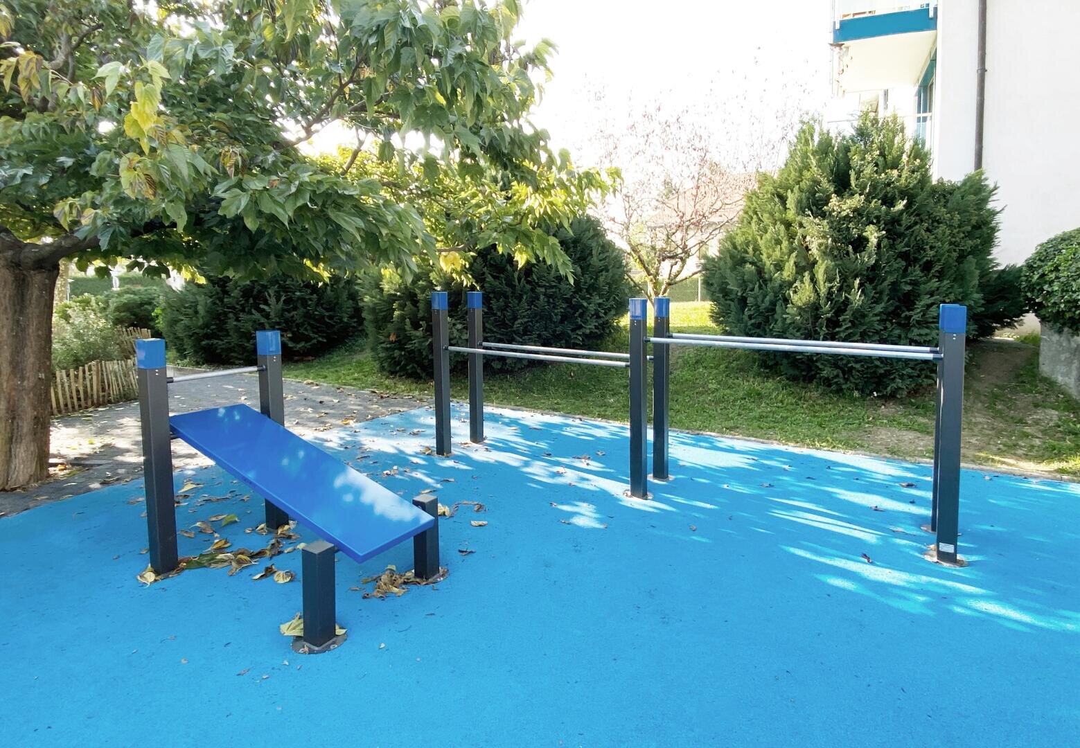 Fitnessbereich im Freien mit blauem Boden, Schrägbank und Parallelbarren. In der Nähe befinden sich Bäume und Sträucher, und im Hintergrund ist ein Gebäude zu sehen. Auf dem Boden liegt Laub verstreut.