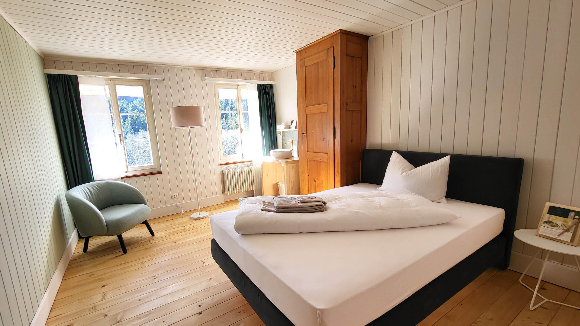 Helles, gemütliches Schlafzimmer mit einem Doppelbett, gefalteten Handtüchern, einem Holzschrank, einem grünen Sessel, einer Stehlampe, einem kleinen runden Beistelltisch und zwei großen Fenstern mit Vorhängen, die natürliches Licht hereinlassen.