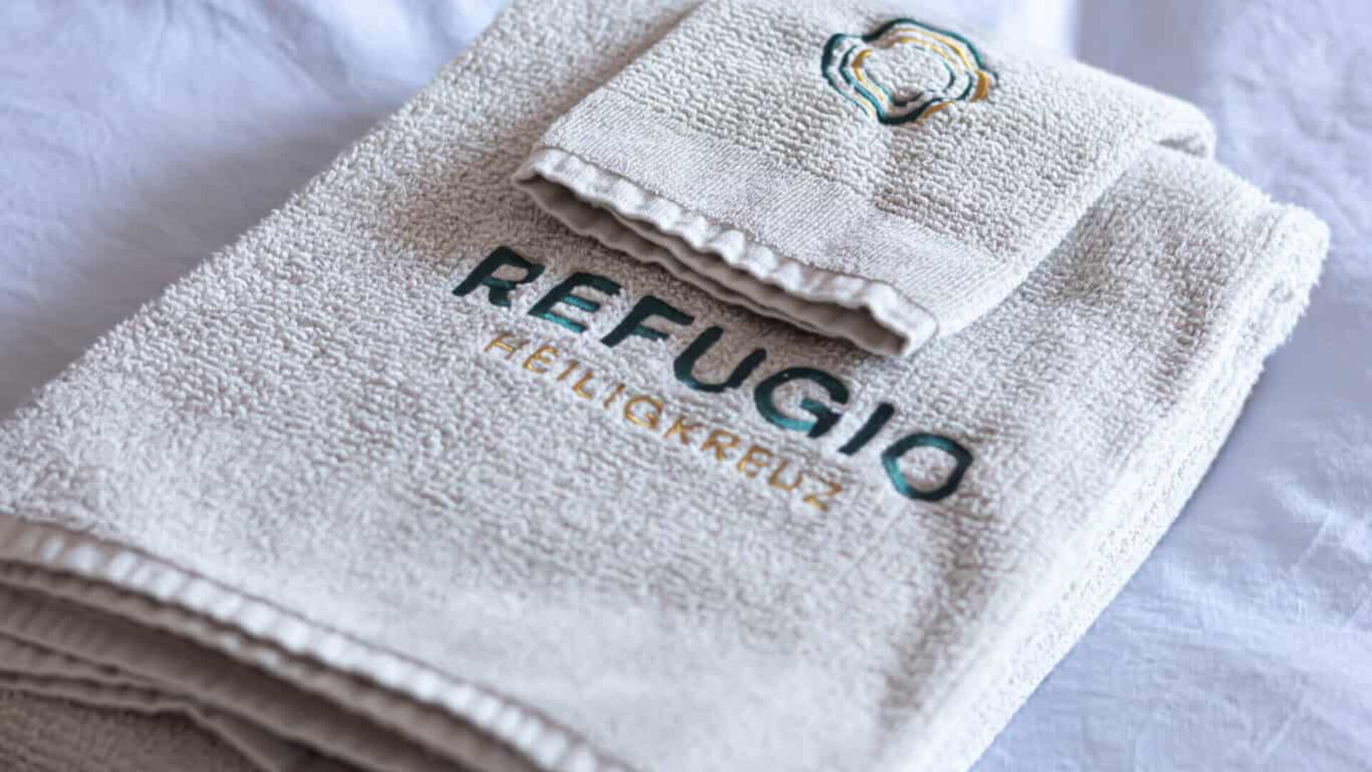 Ein gefaltetes, cremefarbenes Handtuch mit aufgesticktem Wort REFUGIO und HEILIGKREUZ, dazu ein passender Waschlappen mit abstraktem, aufgesticktem Logo, auf weißem Untergrund.