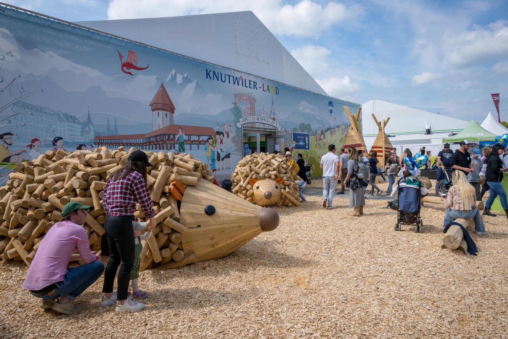 Menschen versammeln sich im Freien in der Nähe grosser hölzerner Igelskulpturen vor einem Zelt mit der Aufschrift "Knutwiler-Land". Kinder spielen und Erwachsene unterhalten sich, während ein Wandgemälde das Zelt ziert und Holzspäne den Boden bedecken.