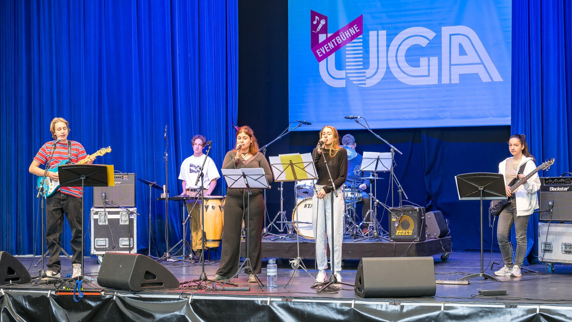 Eine Band von fünf jungen Musikern tritt auf der Bühne mit Gitarren, Schlagzeug und Mikrofonen auf. Hinter ihnen befindet sich ein blauer Vorhang und ein großes Schild mit der Aufschrift UGA EVENT@LINE. Notenständer halten ihre Notenblätter.