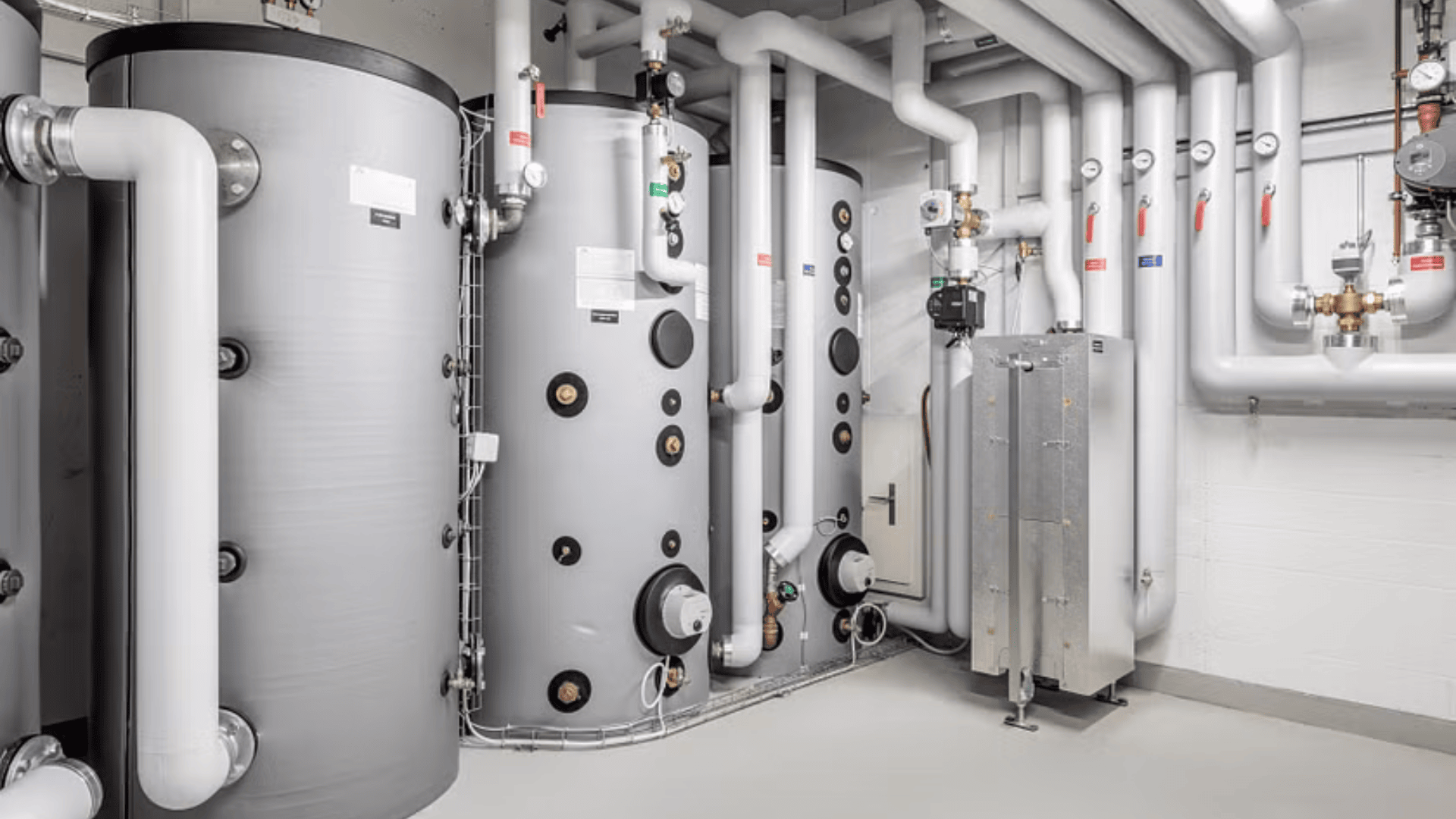 Ein sauberer Kesselraum mit großen zylindrischen Wassertanks und zahlreichen weißen Rohren, die mit an der Wand montierten Ventilen und Messgeräten verbunden sind. Der Boden und die Wände sind weiß, und der Raum wirkt gut organisiert und gepflegt.