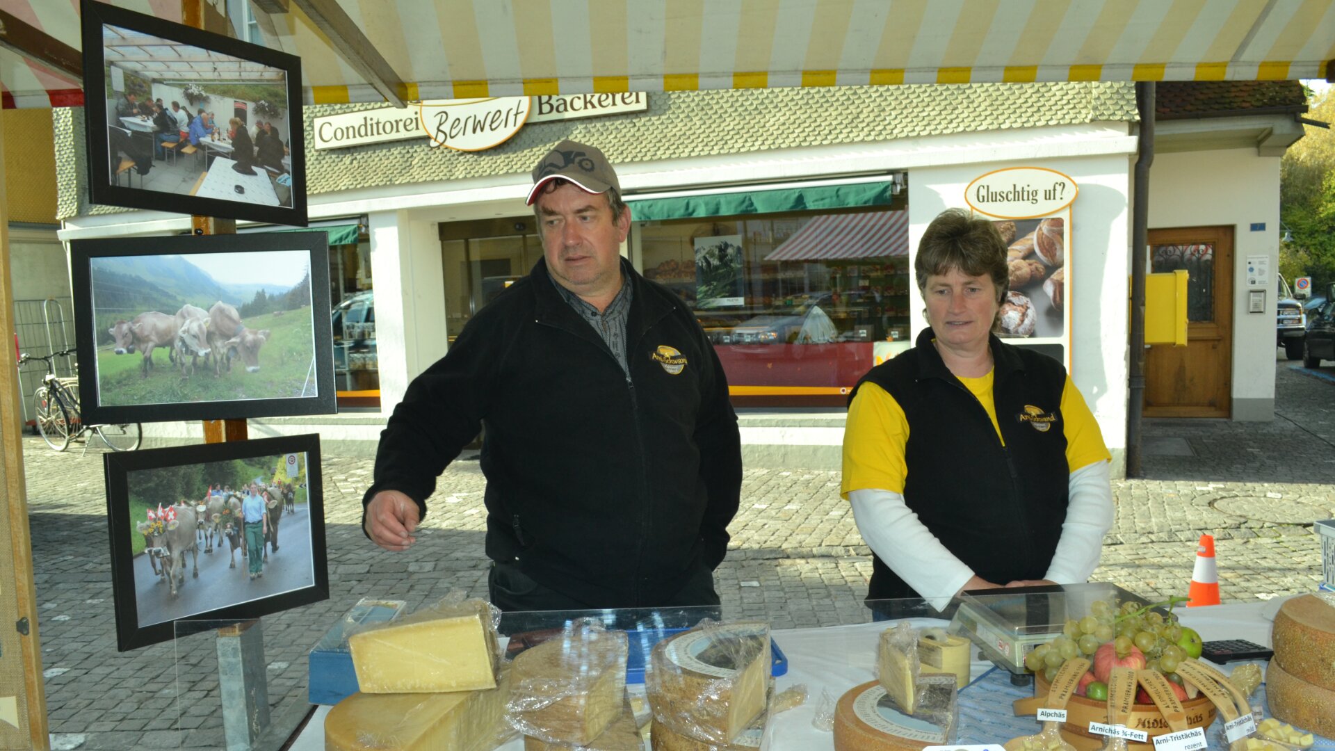 Zwei Marktverkäufer stehen hinter einem Tisch mit verschiedenen Käsesorten und Weintrauben. Hinter ihnen ist das Schaufenster einer Bäckerei zu sehen. An der Seite ihres Standes sind Fotos von Kühen und Kühen beim Hüten zu sehen.