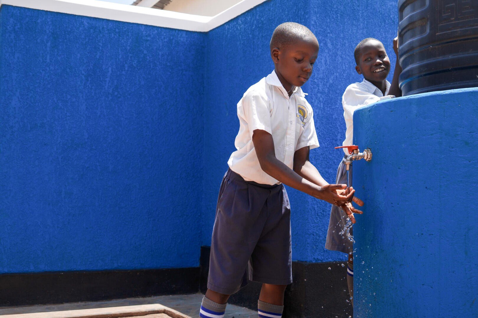 Zwei Schulkinder in Uniformen waschen sich die Hände an einer Wasserstation im Freien mit einer blauen Wand im Hintergrund. Ein Kind benutzt den Wasserhahn, während das andere lächelnd neben dem Wassertank steht.