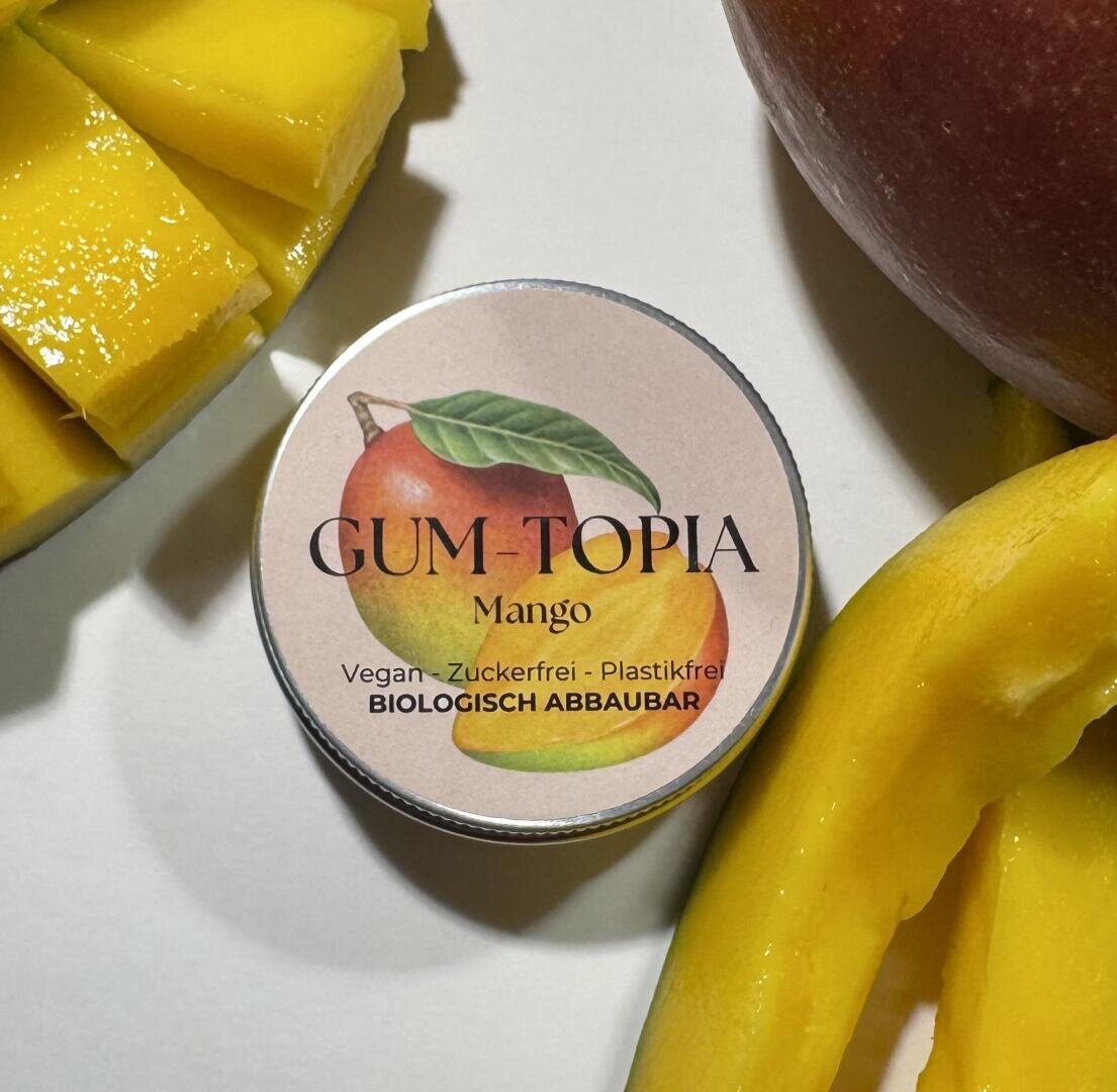 Eine kleine runde Dose mit der Aufschrift GUM-TOPIA Mango steht auf einer weißen Fläche, neben der Mangoscheiben und eine ganze Mango liegen. Das Etikett zeigt eine Mango-Illustration und den Text, dass es vegan, zuckerfrei, plastikfrei und biologisch abbaubar ist.