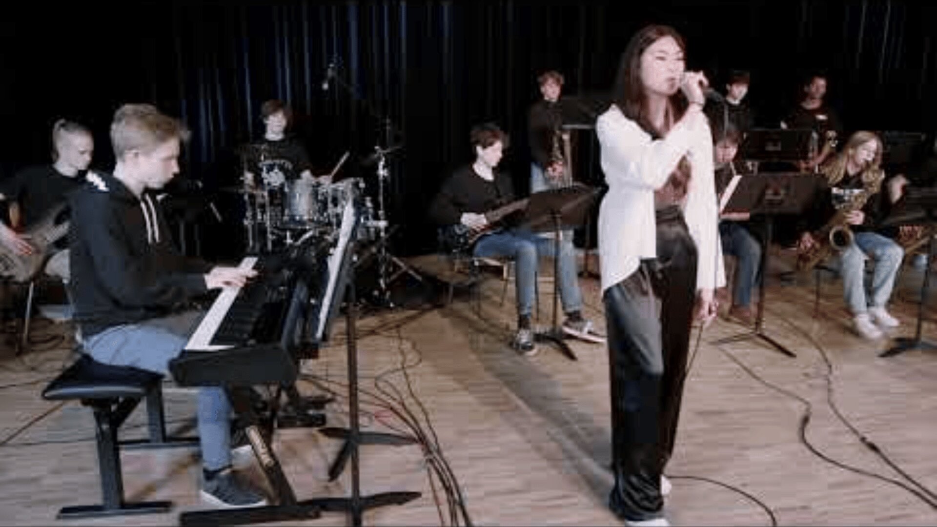 Eine junge Frau singt in ein Mikrofon, während eine Gruppe von Musikern hinter ihr auf einer Bühne mit Holzboden und dunklen Vorhängen Instrumente wie Keyboard, Schlagzeug und Blechbläser spielt.