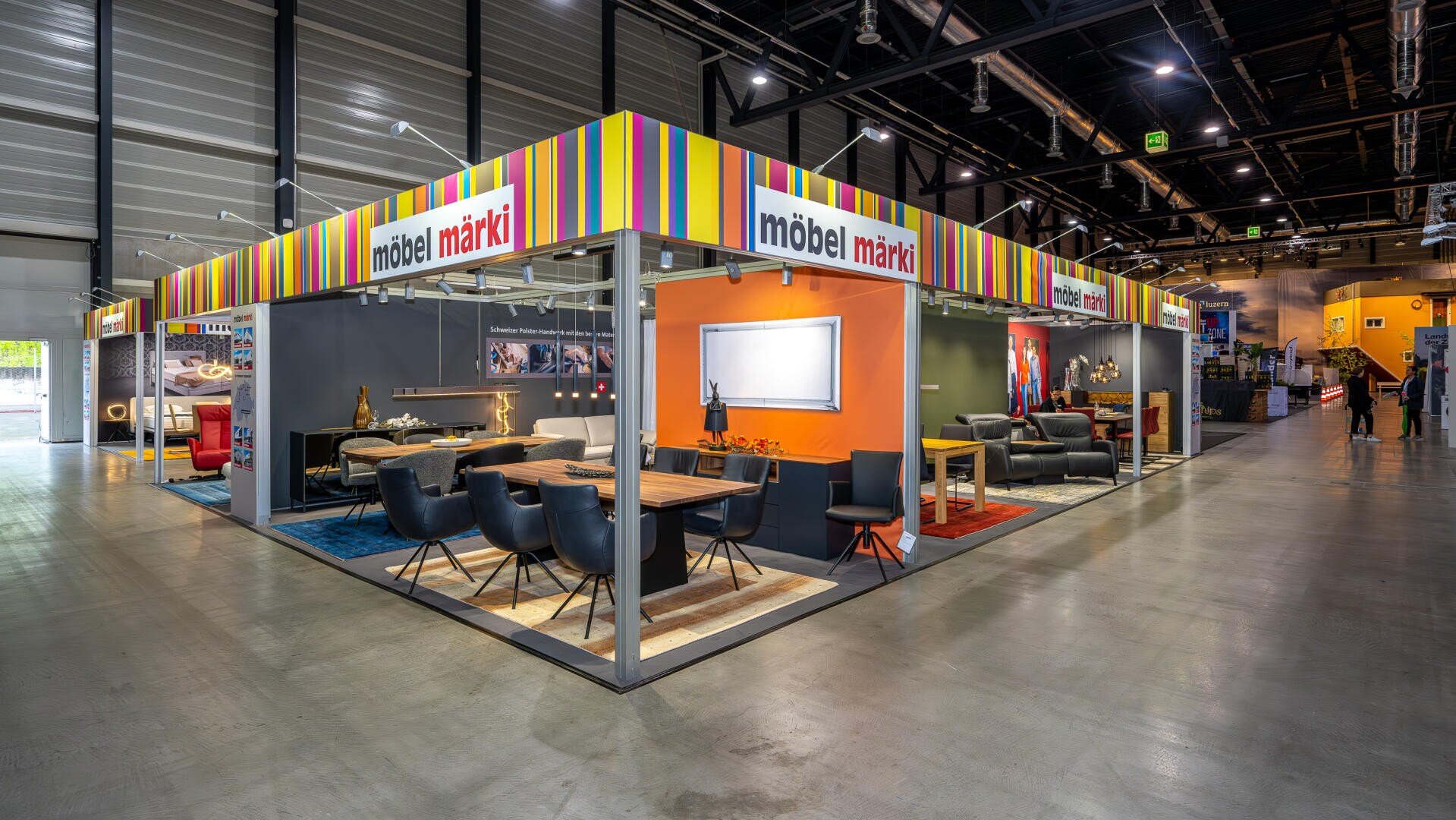 Ein moderner Möbel-Showroom zeigt verschiedene Wohnzimmereinrichtungen mit Sofas, Tischen, Stühlen und Dekoration. Der Stand verfügt über eine bunt gestreifte Beschilderung mit der Aufschrift möbel märki und eine helle Beleuchtung in einem geräumigen Innenbereich.