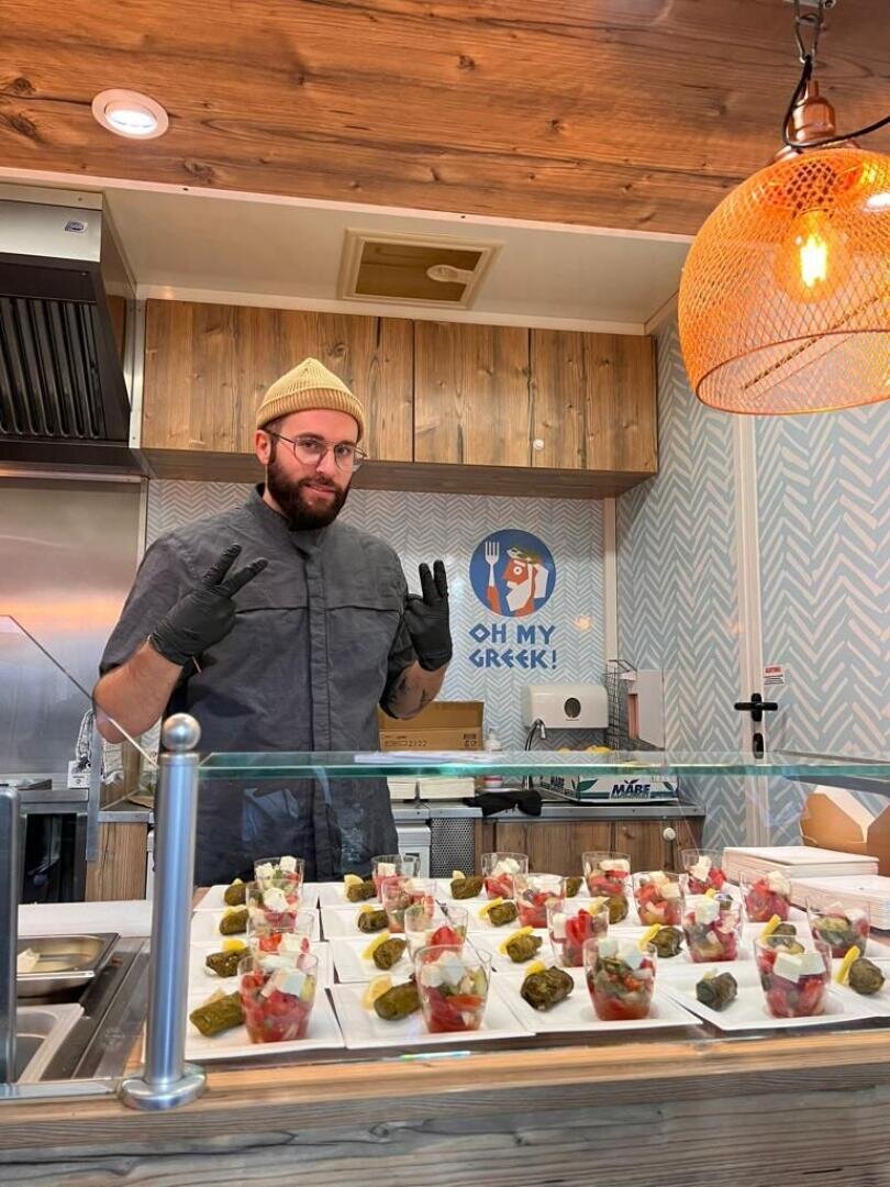 Ein Mann mit gelber Mütze, Brille und schwarzen Handschuhen steht hinter einem Tresen und stellt Teller mit Essen aus. Er macht mit beiden Händen ein Friedenszeichen in einem Restaurant, in dem ein Schild mit der Aufschrift OH MEIN GRIECHISCH! an der Wand hängt.