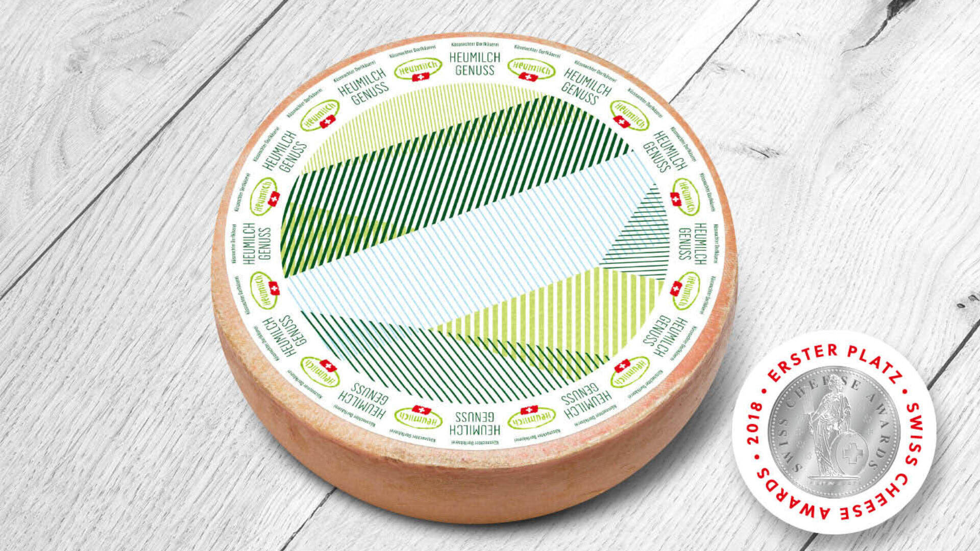 Ein runder Laib Heumilch-Käse mit einer bunten, gestreiften Etikette liegt auf einer Holzfläche. Daneben liegt eine Medaille des Swiss Cheese Awards.